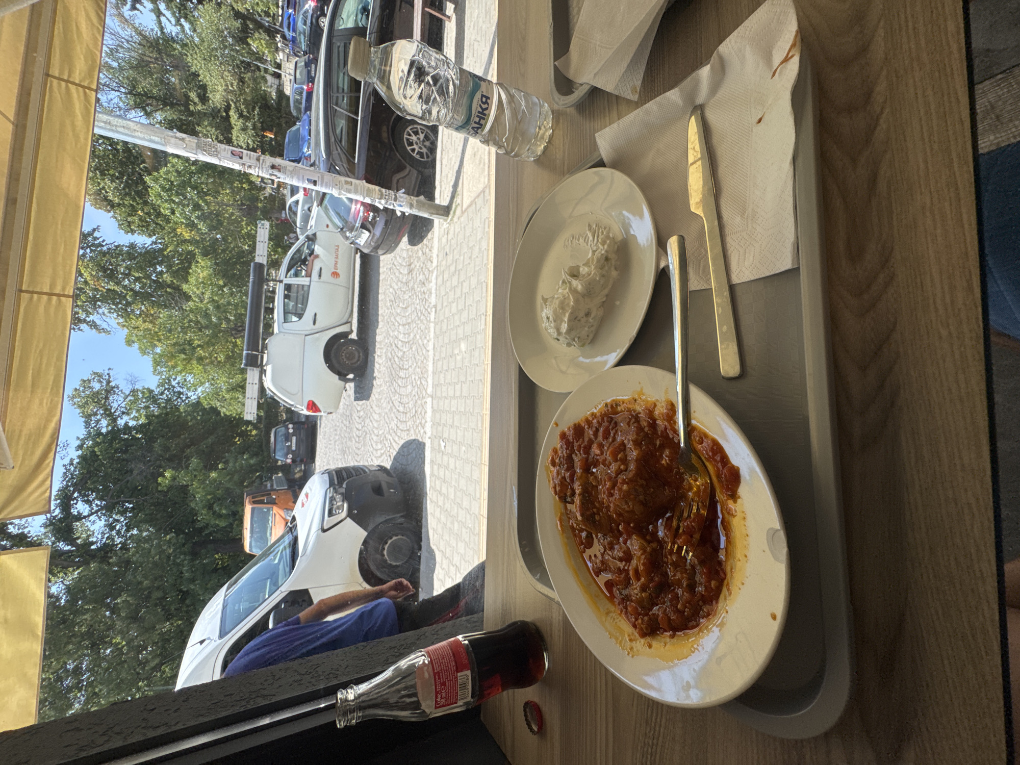 The image shows a table setting with food and drinks at an outdoor establishment, likely a cafe or restaurant, with a view of a street in Sofia, Bulgaria. In the foreground, there is a gray tray holding a white plate with a dish of meatballs in tomato sauce, a fork, and a small white plate with a dip, possibly yogurt-based with herbs. Next to the main plate, a knife rests on a white napkin. A bottle of Coca-Cola and a bottle of water are also on the tray. In the background, through a window or open space, a street scene is visible. Several vehicles are parked or moving on a cobblestone street, including a white van, a small orange truck, a dark SUV, and a white pickup truck with a ladder rack. Trees and a bright blue sky suggest it is daytime and pleasant weather. The establishment appears to be covered by a yellow awning. The overall impression is a casual meal enjoyed outdoors with a view of city life.