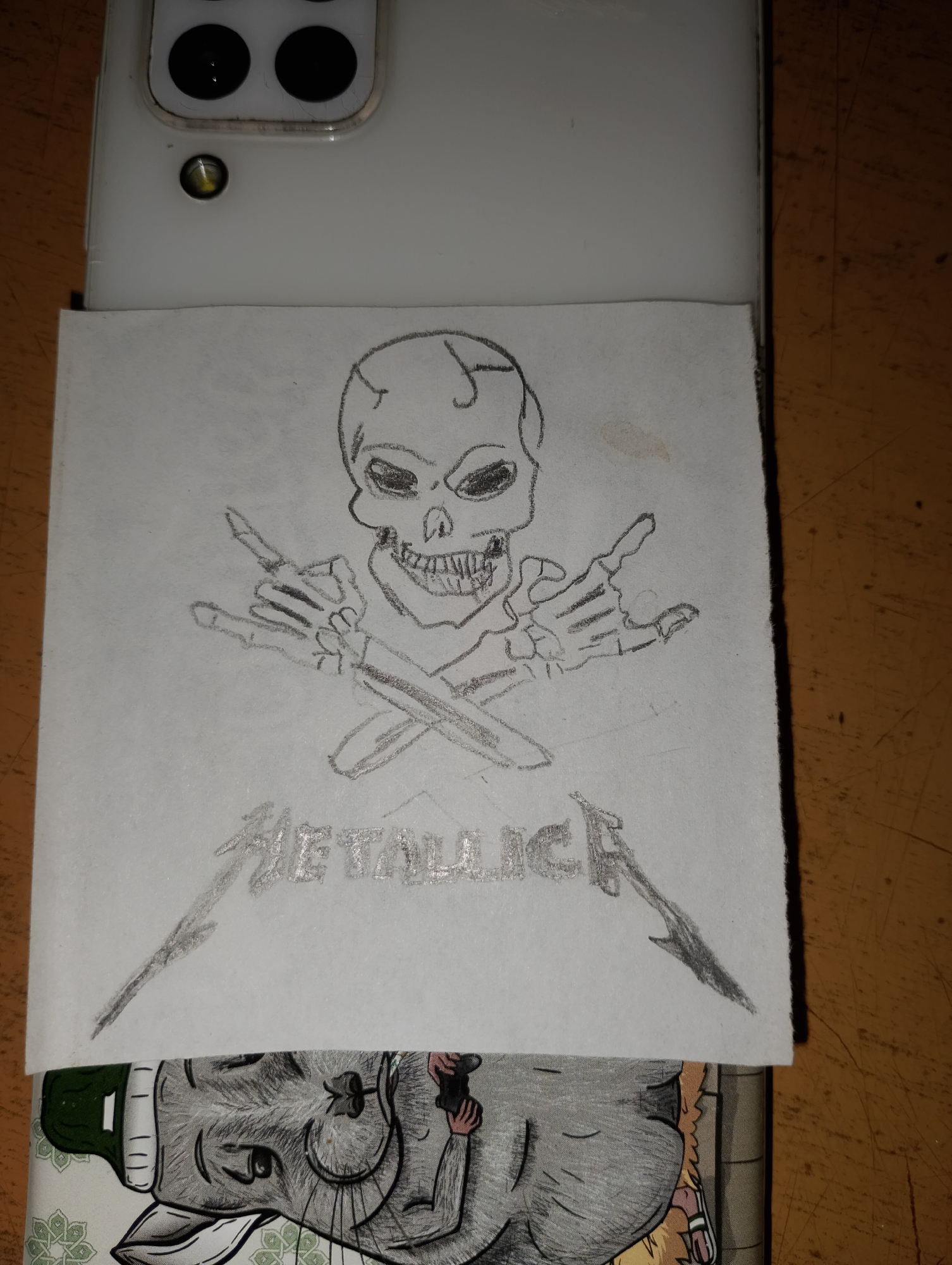 A sketch of a skull with crossed bones and the word "METALLICA" is placed on top of a white smartphone, partially obscuring the camera lenses. The sketch is on a piece of paper, and below it, another drawing of a cat is visible on the back of the phone case. The scene appears to be indoors, with a wooden surface in the background.