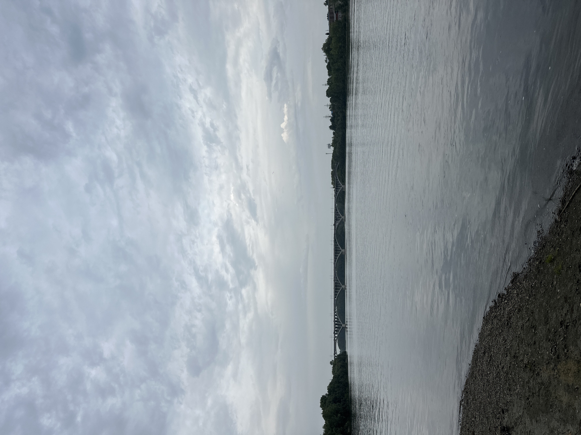 This is an outdoor, full-shot, eye-level photograph of a wide, calm river on an overcast day. A long, multi-arched concrete bridge spans the river in the middle distance. The riverbanks on either side are lined with dense green trees. The foreground features a pebbly and sandy shore. In the far distance, a few buildings and power lines are visible on the right bank. The sky is filled with a thick layer of gray clouds. This scene is likely the Ob River near Biysk, Russia, as the bridge is identifiable as the railway bridge connecting Biysk to the left bank of the Ob.