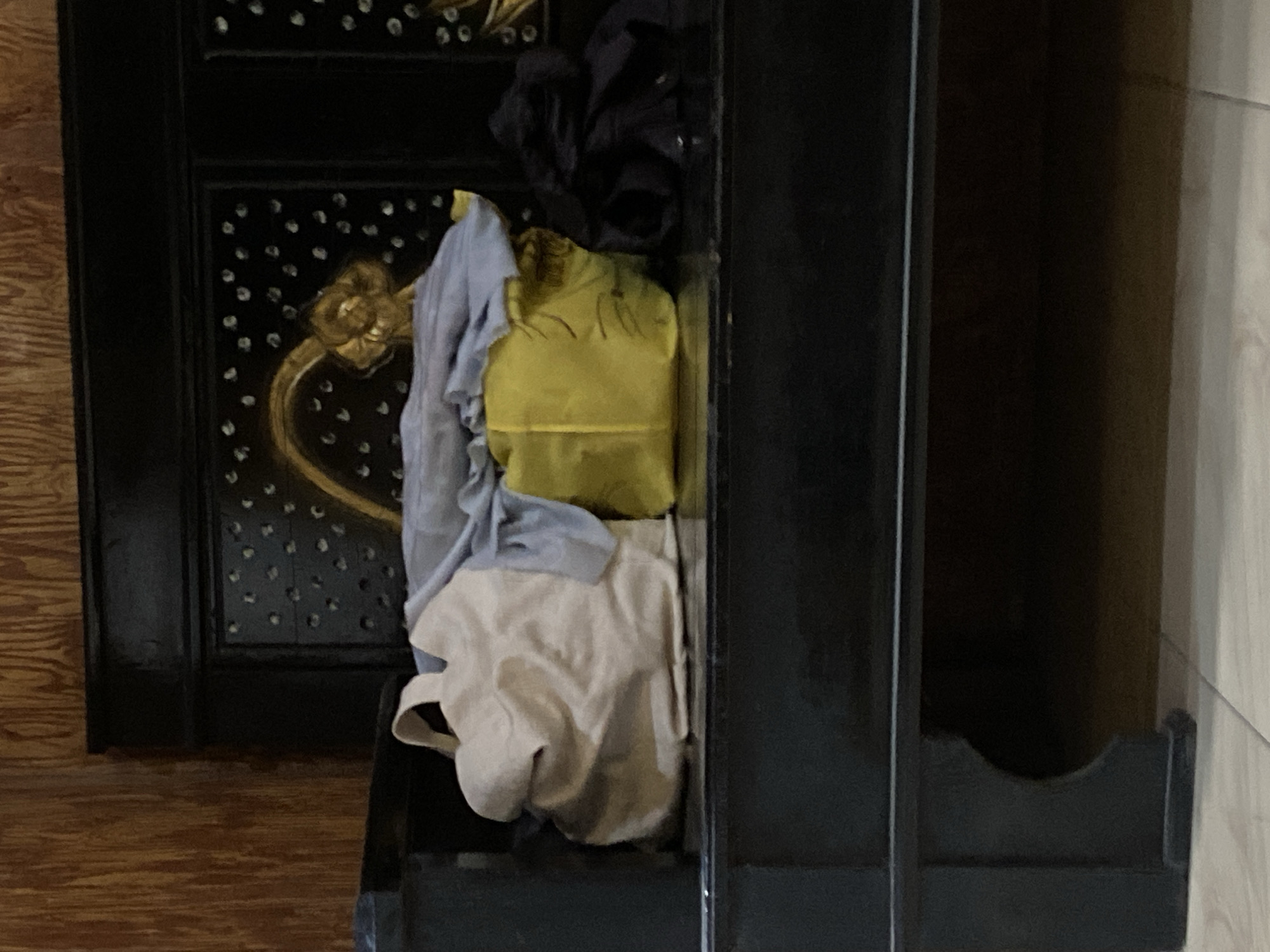 The image shows a black bench with decorative carvings on its backrest. On the bench, there are several items, including a beige tote bag, a yellow bag, and some dark-colored fabric, possibly clothing. The background is a wooden wall with a vertical grain pattern. The floor appears to be tiled. The overall scene suggests a domestic setting, possibly in a home in Plosokandang, Indonesia, where items are stored or placed on a bench. There are no people visible in the image.