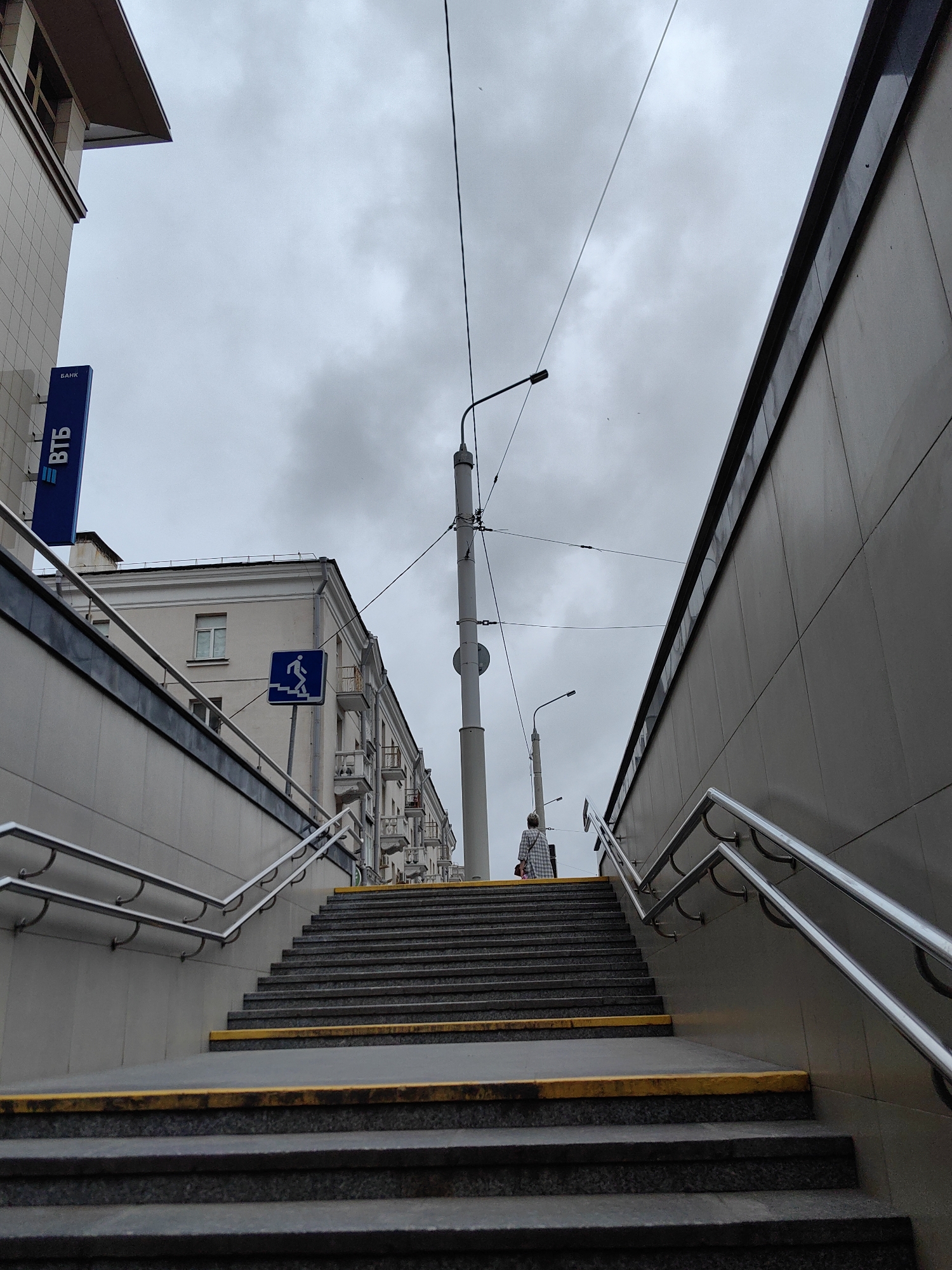 The image shows an outdoor staircase leading down into what appears to be a pedestrian underpass or entrance to a building, likely in Minsk, Belarus. A person, dressed in a plaid shirt and carrying a bag, is walking up the stairs. To the left, a blue sign with white Cyrillic lettering, "БАНК ВТБ", indicating a VTB Bank branch, is visible on the side of a building. Above the staircase, a sign depicting a person walking down stairs is mounted on a pole, indicating a downward path. Overhead, telephone or power lines crisscross the cloudy sky, and streetlights are mounted on poles. The architecture of the buildings suggests an urban environment, with balconies and multiple windows visible on the structures in the background. The overall mood is overcast and grey.