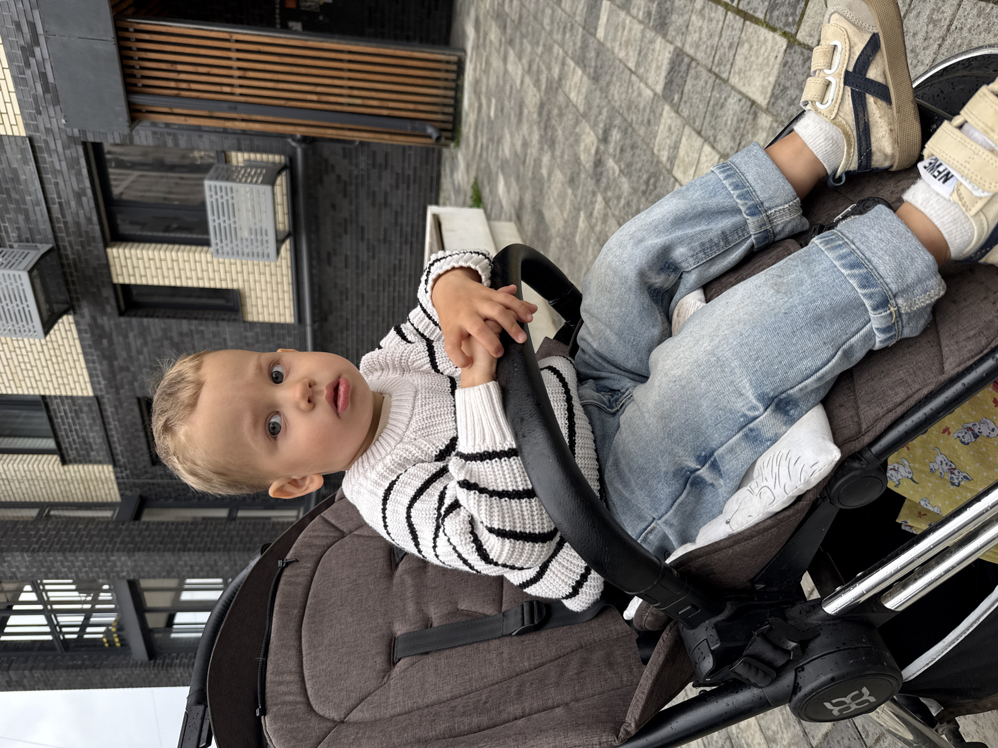 A young boy with blonde hair and blue eyes is sitting in a stroller outside. He is wearing a white sweater with black stripes and light blue jeans. His hands are clasped in front of him. The stroller is dark brown and has a black safety harness. In the background, there is a modern building with dark brick and lighter brick accents. There are also some wooden panels and outdoor seating. The ground is paved with gray and black tiles. The scene appears to be in an urban or suburban outdoor area.