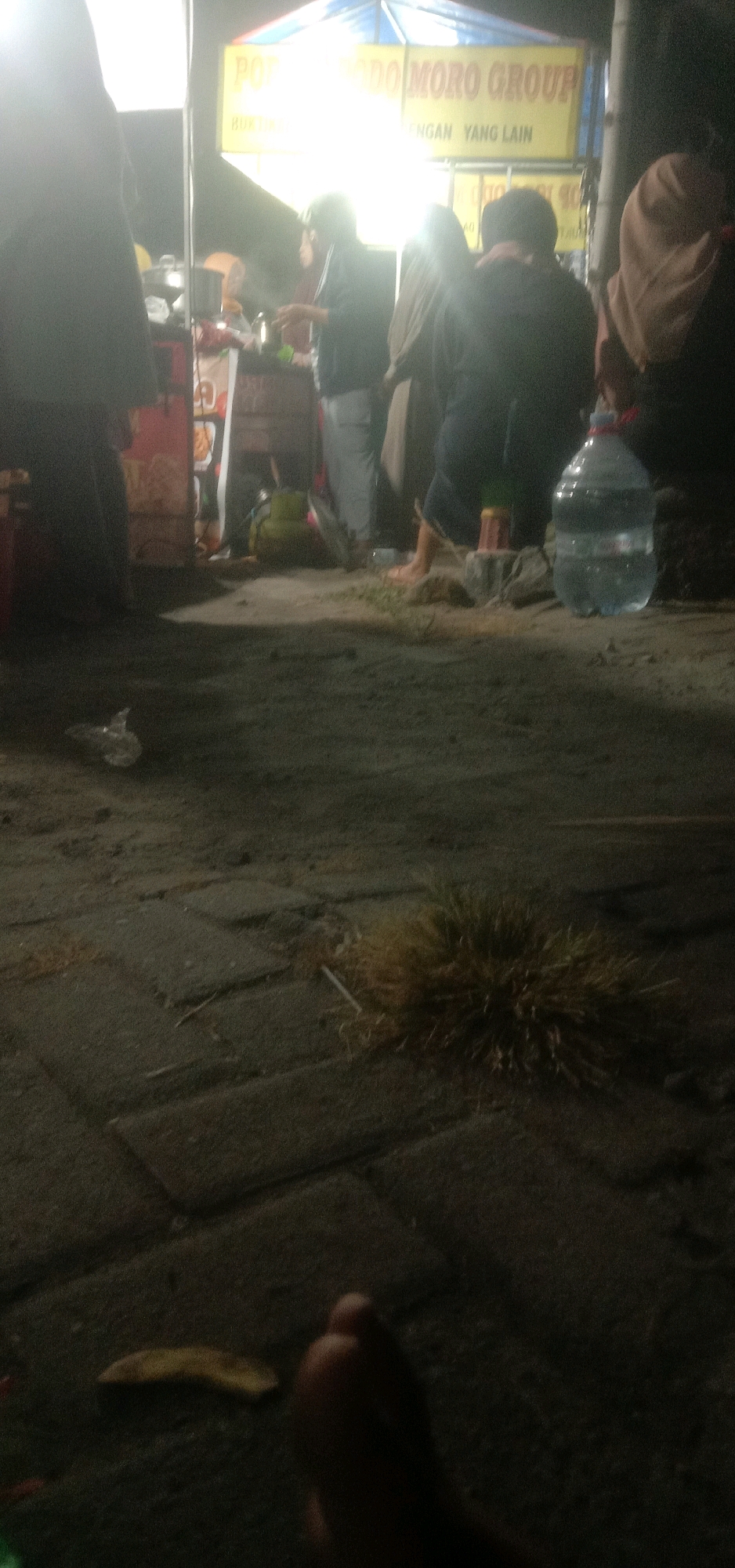The image depicts a dimly lit scene, likely at night, with several people gathered around a food stall. The stall has a yellow banner with text that appears to be in Indonesian. In the foreground, a person's foot is visible, and a piece of fruit or vegetable lies on the ground nearby. A large plastic bottle of water stands to the right. The ground is paved with interlocking stones, and there is some scattered debris and dry vegetation. The overall atmosphere suggests a casual, outdoor gathering or market. The location is identified as Banjar Dowo, Indonesia.