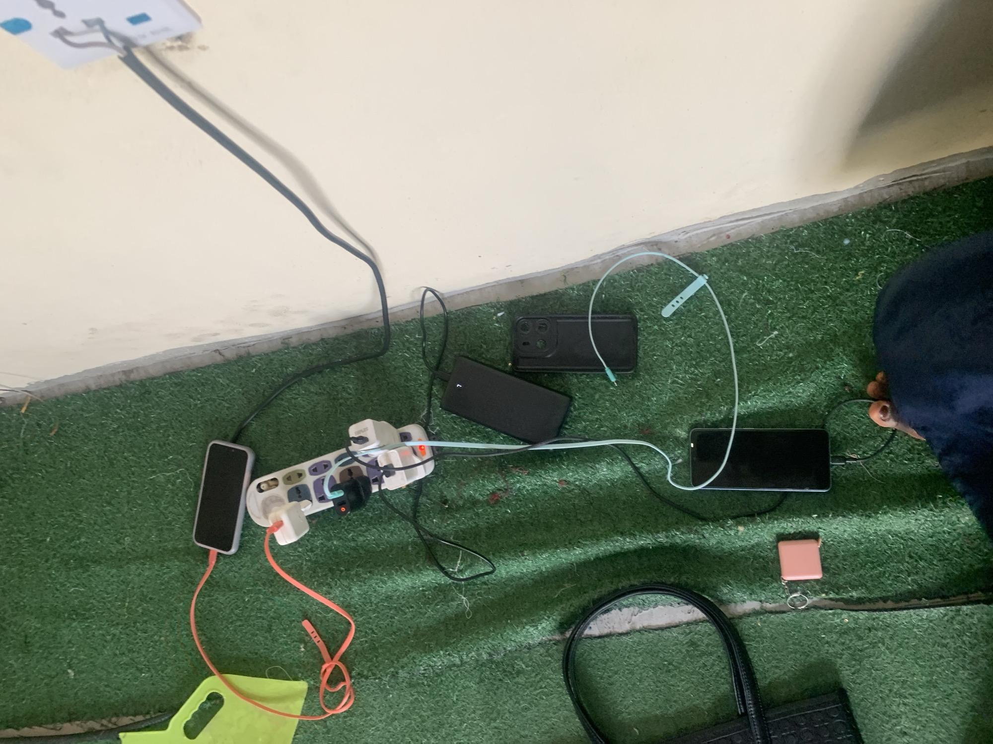In Jauro Yinu, Nigeria, a charging station is set up on a green artificial turf surface. A white multi-socket power strip is plugged into a wall socket, with several charging cables connected to it. Two black smartphones are charging, one of which displays a "7" on its screen. A pink rectangular object with a keyring is also visible on the turf. To the left, a black handbag rests on the ground, and a neon yellow object, possibly a tool or a piece of equipment, is partially visible. A person's foot, wearing dark clothing, is in the lower right corner of the image. The overall scene suggests a temporary or improvised charging area within a residential or public space.