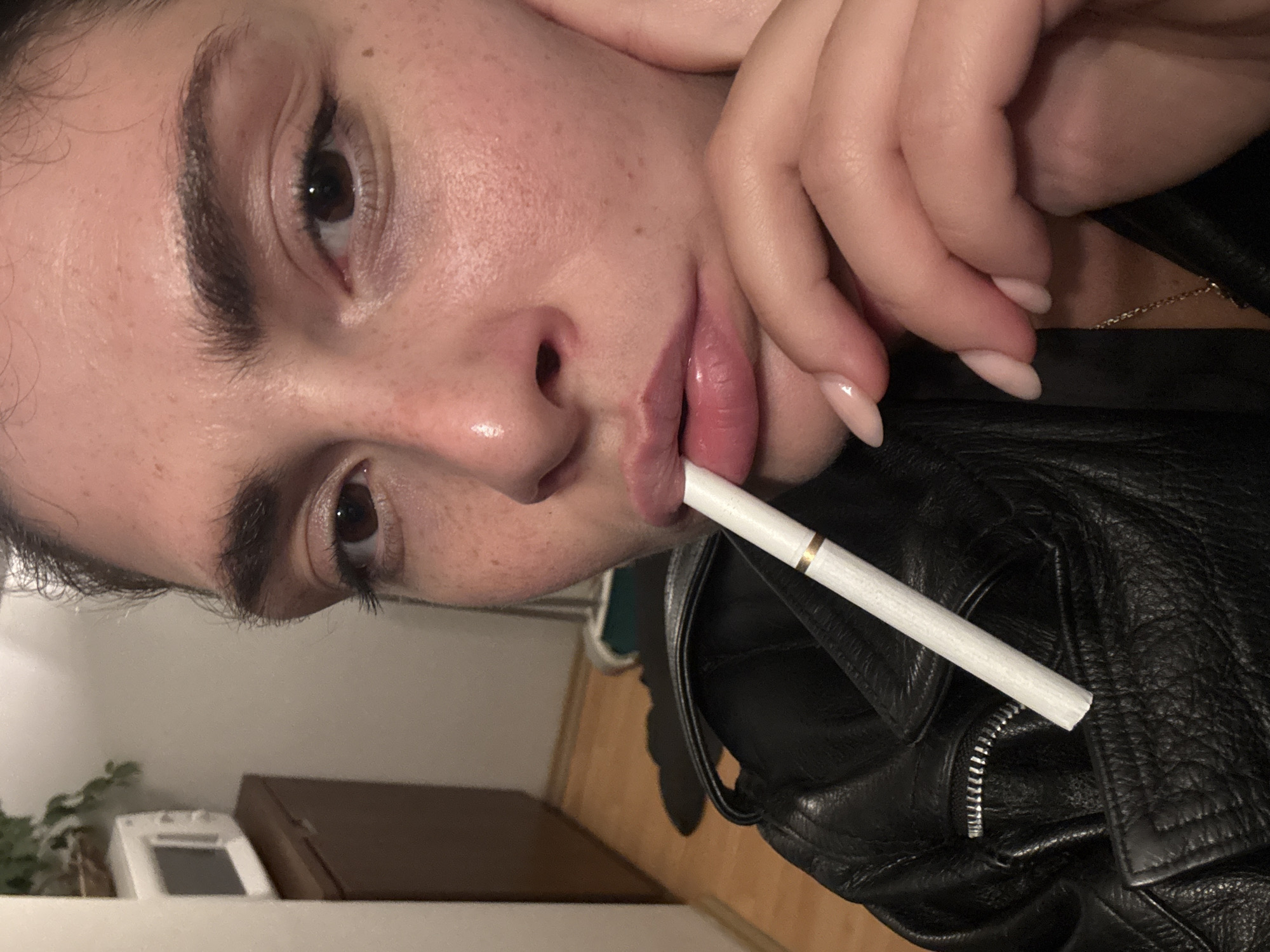 A close-up, angled shot features a person, likely female, with an unlit white cigarette held between their lips. The cigarette has a gold band near the filter. The person's face shows brown eyes, dark eyebrows, and visible freckles on the cheeks and nose. Their lips are full and appear to have a glossy, pink-toned product. A hand with manicured, light pink/nude nails is positioned near the face, resting by the chin area.
The person is wearing a black leather jacket with visible texture and a metal zipper. The background indicates an indoor setting, dimly lit, with a light-colored wall, a dark wooden dresser or cabinet, a small green plant, and a white object resembling a remote control or thermostat visible in the lower part of the frame. The lighting is low and artificial, creating a casual, intimate atmosphere. No explicit location cues are visible within the image to confirm Sofia, Bulgaria, but this is the provided context. There is no visible text.