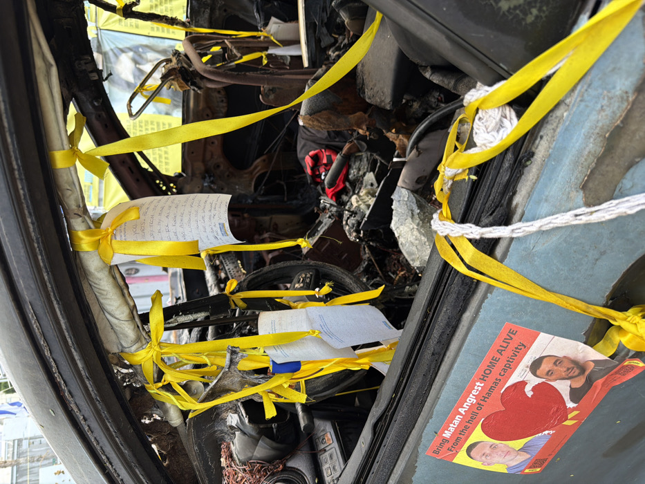 The image depicts the severely burnt interior of a passenger vehicle, likely a sedan, at Hostages Square (Kikar HaHatufim) in Tel Aviv, Israel. The car's dashboard, steering wheel area, and front seating are gutted, showing charred remnants of plastic and metal, indicating extensive fire damage. Numerous yellow ribbons are tied throughout the exposed framework of the vehicle's interior and along the door frame, symbolizing solidarity with hostages. Several handwritten notes are attached to these ribbons; one visible note includes phrases like "BE SAFE," "WE ALL LOVE YOU," and "IM ADMIRE YOU," along with a Ukrainian flag symbol. Affixed to the car's exterior, just below the window line, is a prominent red and white sticker. The sticker features portraits of two men and a broken red heart, with text that reads: "Bring Matan Angrest HOME ALIVE From the hell of Hamas captivity." In the blurred background, additional yellow banners or flags are visible, consistent with a protest or memorial display, and a partial Israeli flag can be seen in the upper left. The scene is outdoors, under daylight conditions.