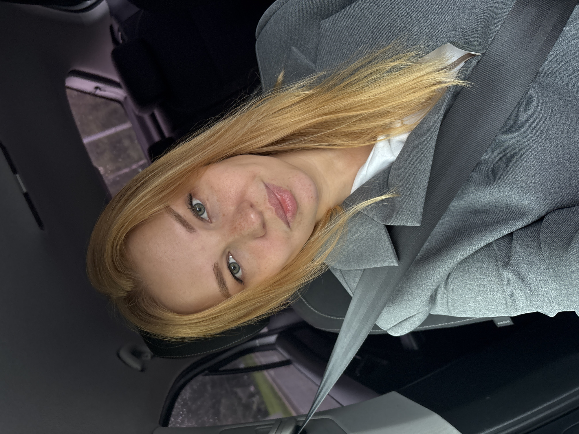 A young woman with blonde hair and freckles is sitting in the passenger seat of a car. She is wearing a gray blazer and a white shirt. A gray seatbelt is fastened across her chest. The background shows the interior of the car and a blurred view of the outdoors through the window. The location is stated to be Rusanovo, Russia.