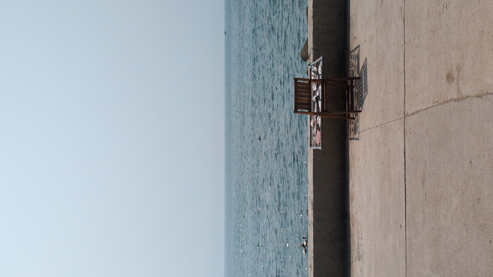 A horizontal, eye-level outdoor shot shows a concrete pier next to a body of water under a clear sky. The sky is light blue. The water has a shimmering, rippled surface. On the pier, a rectangular, brown wooden structure sits. Beside it is a drying rack with clothes. The pier is made of concrete, divided by lines running parallel to the water. The scene is located in the city of Phan Rang – Tháp Chàm, Vietnam. There are no people in the image.