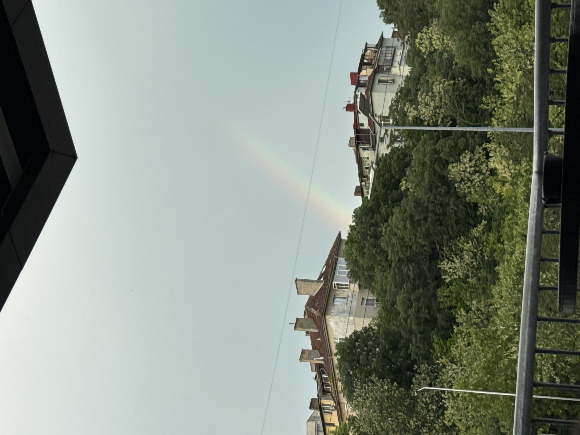 The image shows a rainbow arcing over buildings and trees in what appears to be a residential area. The sky is light gray. On the left side of the image, part of a dark-colored building is visible. The buildings in the middle appear to be apartment buildings with red tile roofs, interspersed with green trees. On the right side of the image, there's a railing. Given the architectural style and vegetation, the scene is likely in Varna, Bulgaria. There are no people visible in the image.