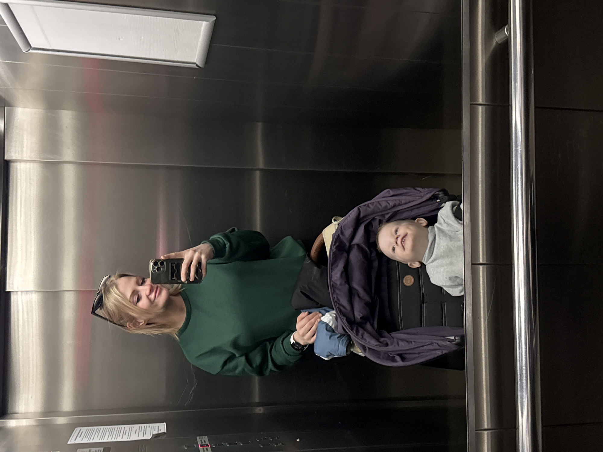 A woman with blonde hair, sunglasses perched on her head, is taking a selfie with a smartphone in a silver elevator. She's wearing a green sweatshirt and holding a baby wrapped in a dark purple blanket. The baby is lying in a black stroller. The elevator has stainless steel walls and a grab bar. There are elevator buttons and a light fixture visible. The location is Saint Petersburg, Russia.