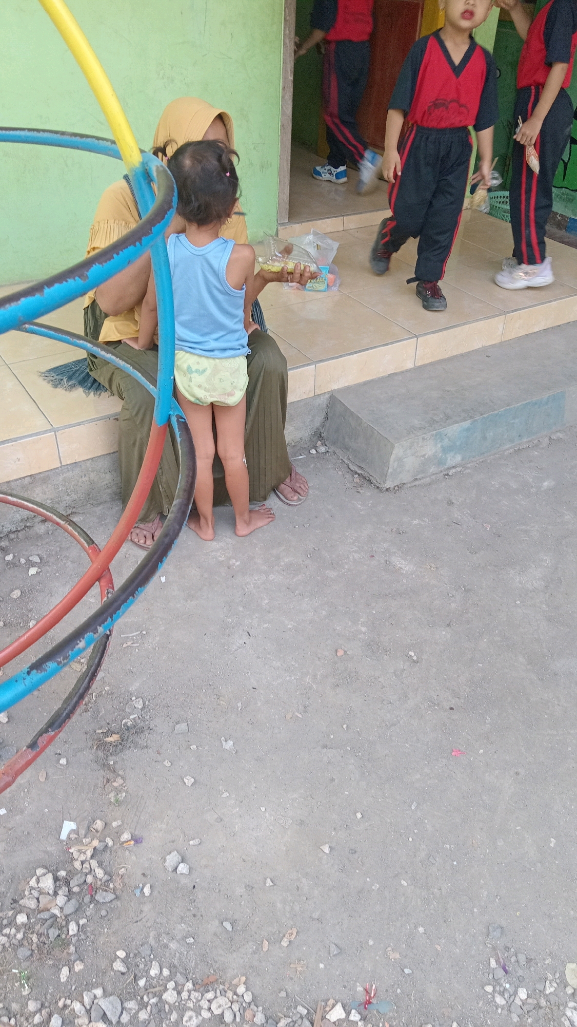 In Kedungwaru, Indonesia, a young child wearing a blue tank top and yellow diaper shorts sits on the lap of a woman wearing a yellow hijab and olive-green pants. The child is holding a clear plastic container with food. Behind them, two other children, one wearing a red and black uniform and the other partially visible in a similar uniform, stand near a doorway. The scene takes place outdoors on a tiled surface adjacent to a light green wall and a concrete step. A colorful metal playground structure is in the foreground, and the ground is covered in loose gravel and debris.