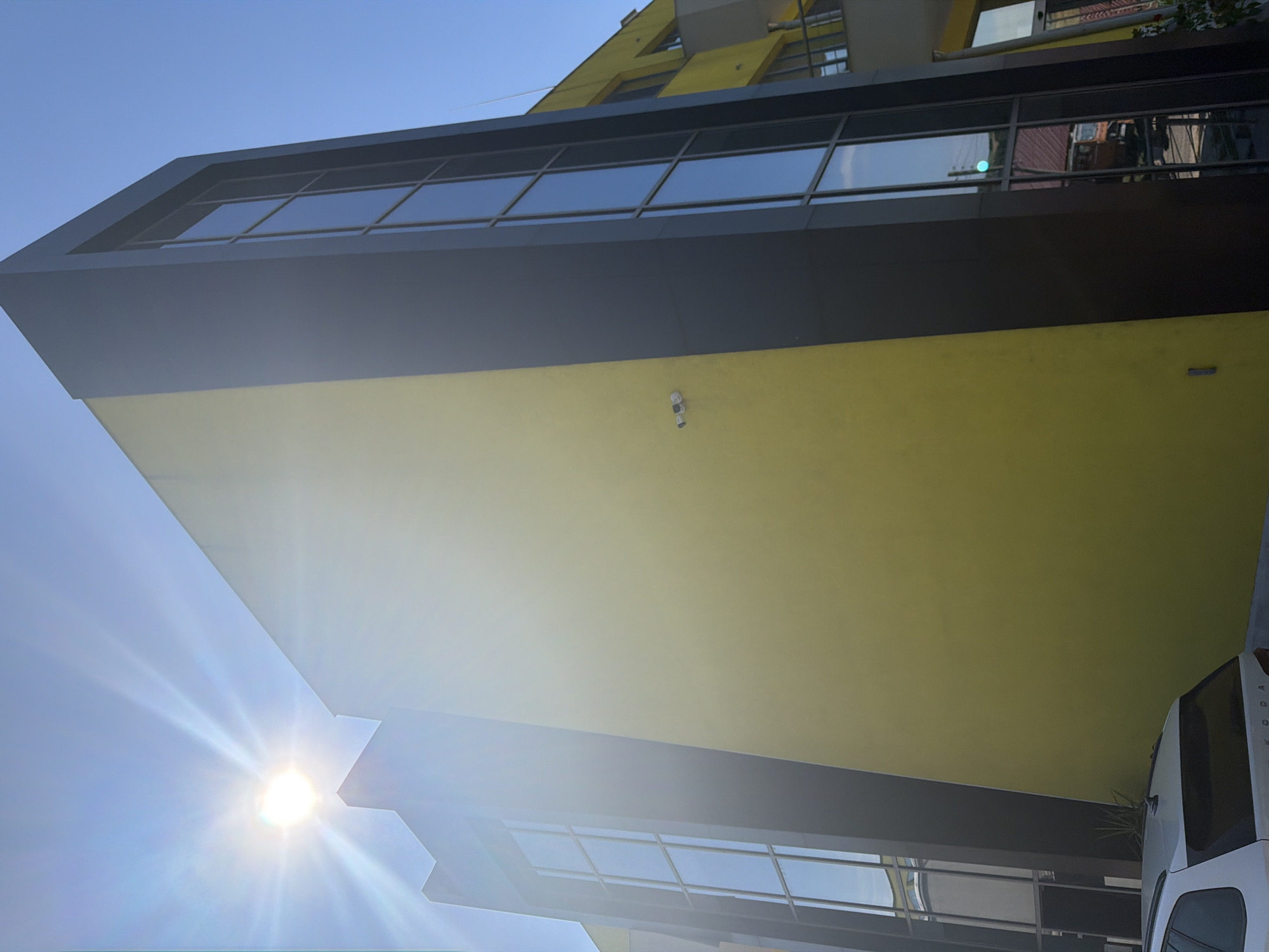 The image captures a modern building with a bright yellow facade and dark gray architectural elements, including tall, narrow windows. The sun is visible in the upper left corner, casting a lens flare across the sky and part of the building. A security camera is mounted on the yellow wall. In the foreground, a white car is partially visible, and a small plant grows near the base of the building. The building is located in the city of Dolno Ablanovo, Bulgaria. The scene is set against a clear blue sky on a sunny day.
