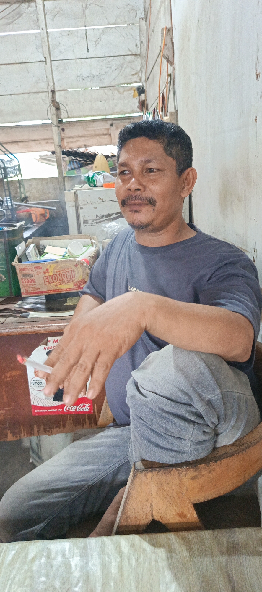 A man with dark hair, a mustache, and a goatee is sitting in a chair, holding a cigarette. He is wearing a dark grey t-shirt and light grey pants. The man is in an interior setting with a wooden table in front of him, on which a box of soap and a Coca-Cola box are placed. The walls are a light color, and there are exposed wooden beams and some items on shelves in the background. The overall environment appears to be modest and possibly a workshop or storage area. The image was taken in Garut, Indonesia.