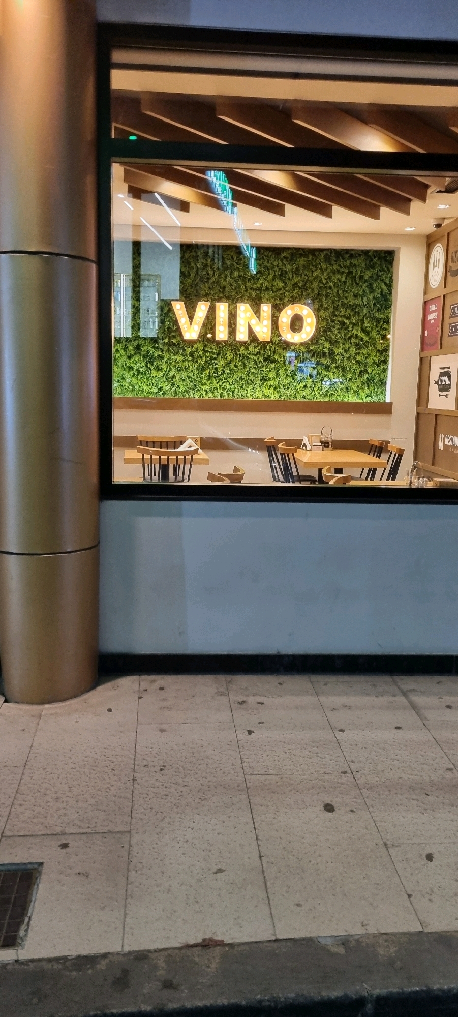 The image shows the exterior of a restaurant named "VINO" in Bashkia Durrës, Albania. The restaurant's name is displayed in illuminated marquee-style letters against a backdrop of artificial green foliage. Inside, through the window, several tables and chairs are visible. Some tables have chairs around them, while others appear to have chairs stacked or removed. Boxes with various labels, including "RESTAURANT," "GRILL HOUSE," and "MENU," are stacked on the right side of the image, suggesting a commercial establishment. The exterior features a large, cylindrical golden pillar on the left and a tiled sidewalk in the foreground. The lighting suggests it is nighttime or dusk.