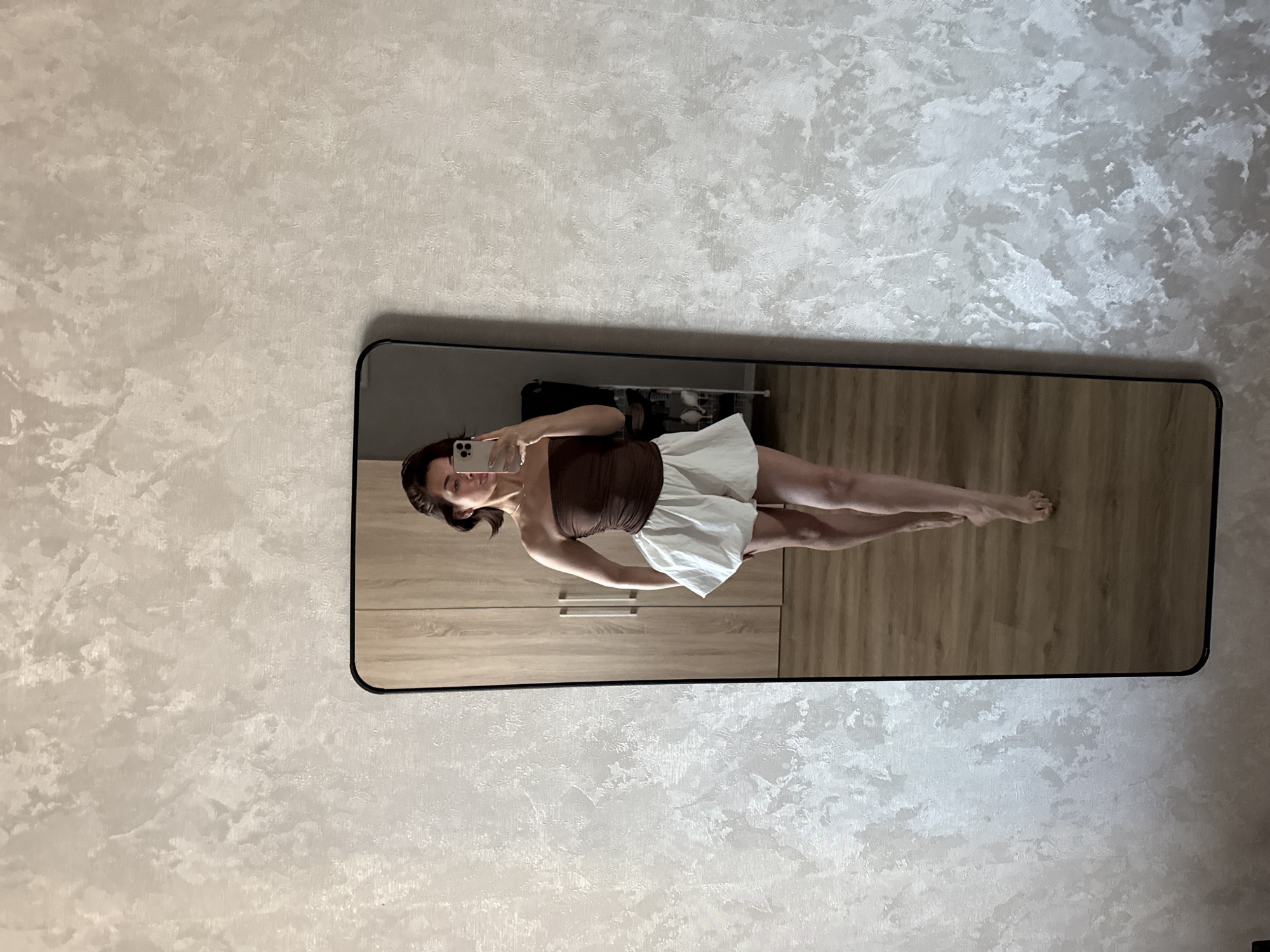 A young woman is reflected in a rectangular mirror with a black frame. The mirror is positioned against a wall with a textured, marbled pattern in light beige and gray tones. The reflection shows the woman holding a smartphone, presumably taking a selfie. She wears a brown strapless top and a white skirt. The background within the mirror includes light wood-grain cabinets with silver handles, a beige wood floor, and a glimpse of a black storage unit containing what appears to be a wire basket. The woman appears to be in an indoor space, possibly a bedroom or dressing area. The location is likely in Лобаново, Russia.