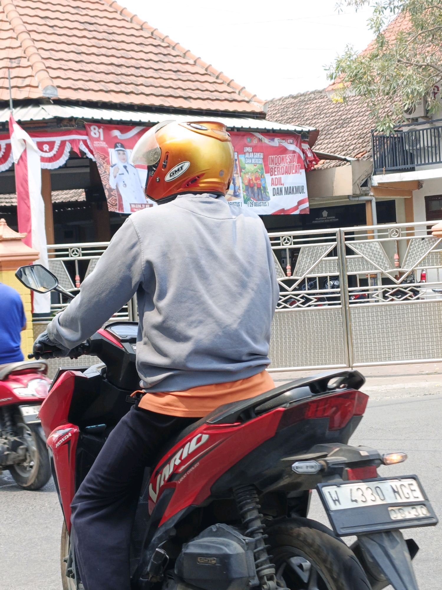 In the city of Ketegan, Indonesia, a person is riding a red Honda Vario scooter. The rider is wearing a gold and orange helmet and a grey long-sleeved shirt over an orange undershirt, with dark blue pants. They are wearing grey gloves and holding the scooter's handlebars. The scooter has a license plate that reads "W 4330 NGE". In the background, a building with a tiled roof is visible, along with a metal fence and banners celebrating Indonesia's independence. One banner features text that translates to "Indonesia, Just, Adorable, and Prosperous." Another banner displays the number "81." There is also a red and white Indonesian flag.