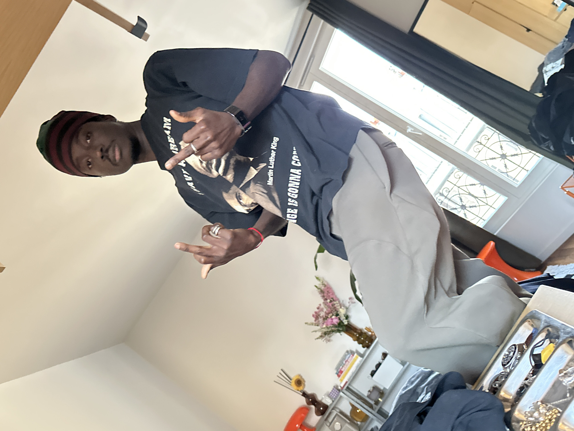 A person stands inside a building. The person is wearing a black t-shirt with an image of Martin Luther King Jr. and khaki pants, and a hat. The person is making hand gestures. In the background, there is a window and a shelf with flowers and other objects. The image was taken in Saint-Ouen-sur-Seine, France.
