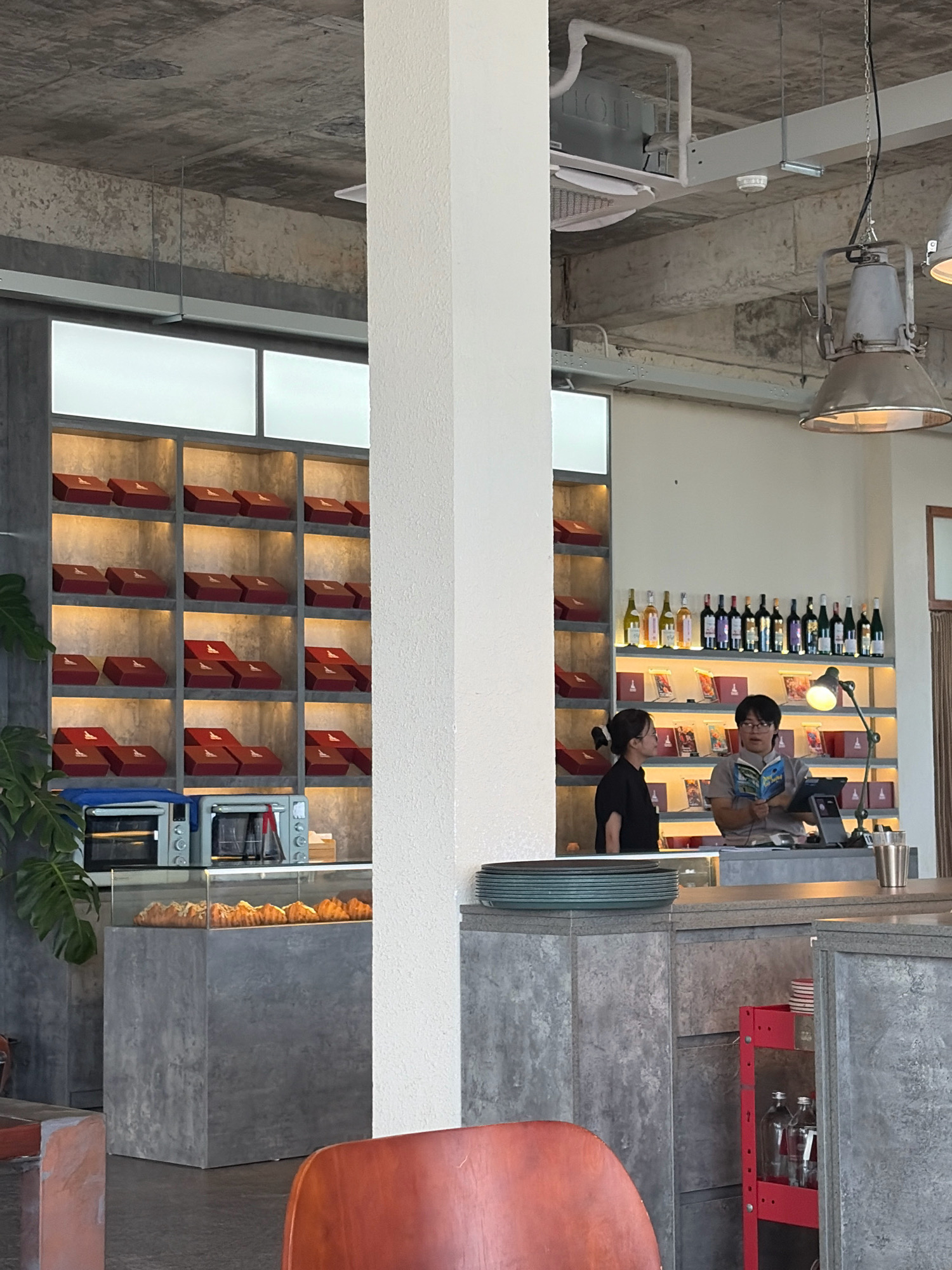 This is a wide shot of a cafe interior in Da Nang, Vietnam. The cafe features an industrial design with exposed concrete ceilings, grey concrete walls and counters, and industrial pendant lights. On the left, shelves are filled with red gift boxes. Below the shelves are two silver mini ovens and a display case containing baked goods. A large white pillar divides the space. On the right, a counter area features two people. A woman in a black uniform stands to the left, looking at a man in a grey shirt who is reading a book. Behind them, shelves display bottles of wine and more red gift boxes. The overall atmosphere is modern and minimalist.