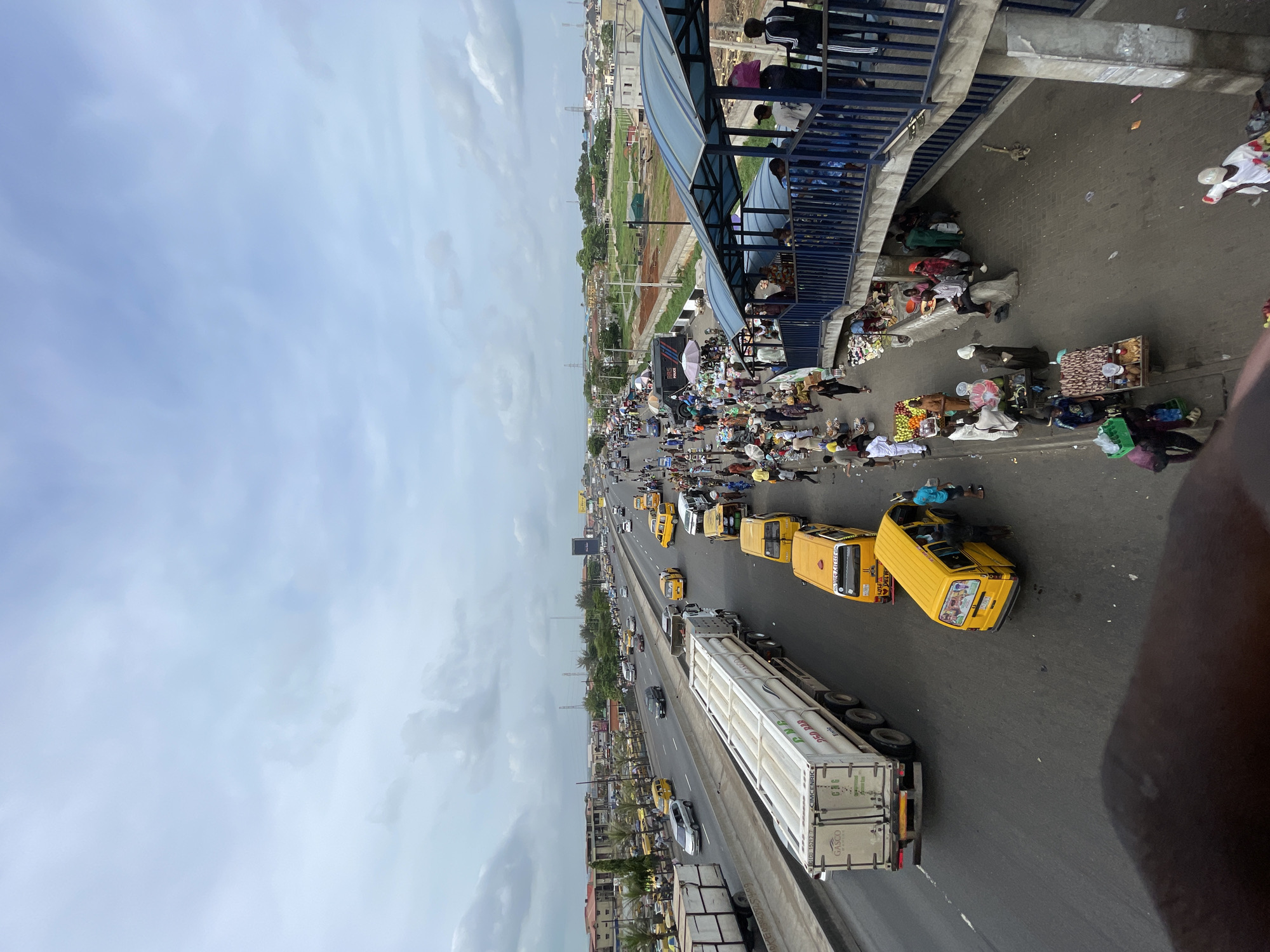 The image shows a busy road in Isheri, Nigeria, viewed from an elevated position, likely a pedestrian bridge. The road is filled with traffic, including yellow buses (danfos), cars, and a large white semi-trailer truck. There are people on the pedestrian bridge, some looking down at the scene below. Alongside the road, there are informal vendors and pedestrians, creating a crowded marketplace atmosphere. The sky is overcast. There is evidence of construction or landscaping in the background.