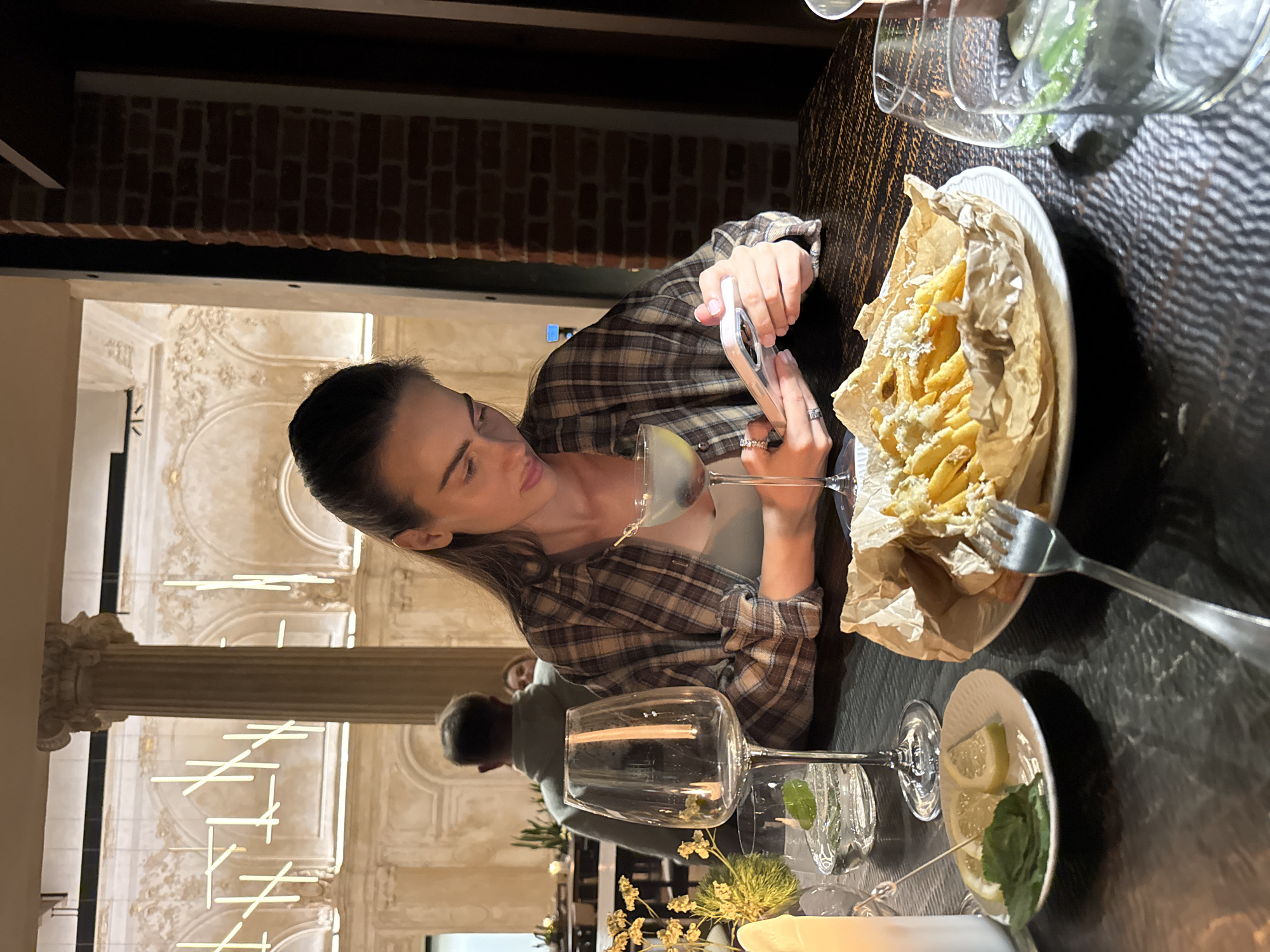 An indoor restaurant or bar scene is depicted, situated in the city of Unknown, Unknown, characterized by a blend of classical architecture and modern design. The environment is dimly lit with warm ambient light.
In the foreground, a young adult female with dark hair, pulled back, is seated at a dark, textured table. She wears a light-colored undergarment beneath an open brown, beige, and white plaid button-down shirt. Her hands are engaged with a light-colored smartphone, on which her gaze is fixed. A ring is visible on her left ring finger. Directly in front of her is a stemmed cocktail glass containing a pale liquid and a dark garnish on a pick. To her left, an empty stemmed wine glass and a rock glass with water, ice, and a mint leaf are visible. A small white plate with two lemon wedges and fresh mint leaves, skewered by a cocktail pick, rests beside an unlit white candle. To her right, a plate holds a serving of french fries presented in crumpled brown parchment paper, topped with a white, crumbled ingredient, and a silver fork resting on the side. Another rock glass with water, ice, and mint leaves is partially visible on the far right.
The background reveals a spacious interior featuring high ceilings, large light-colored walls adorned with ornate plasterwork, and multiple arched alcoves. Tall classical columns with intricate capitals are prominent. Modern linear LED tube lights are suspended from the ceiling at various angles, casting light throughout the space. Further back, a male figure wearing a light grey hooded sweatshirt is seated with his back partially to the camera at another table. A blurred face of an additional person is faintly visible behind him. A section of a red brick wall is also visible on the right side of the background, contrasting with the classical elements. Dark furniture is visible in the distance.