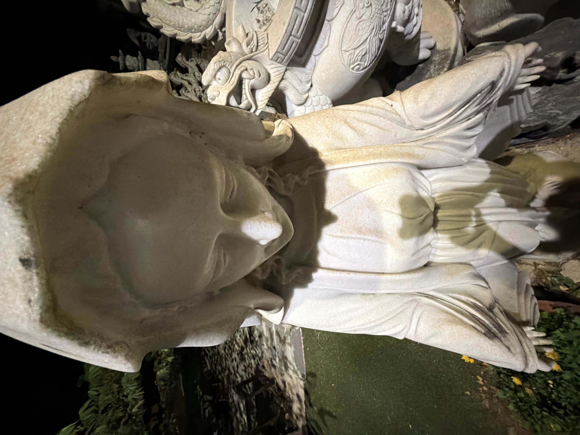 A marble statue of a hooded figure is positioned in a garden setting in Đà Nẵng, Vietnam. The figure is seated, with hands outstretched. A stone dragon sculpture is visible in the background. The statue is illuminated by artificial lighting. The environment includes greenery and possible stone elements. The scene appears to be set at night.