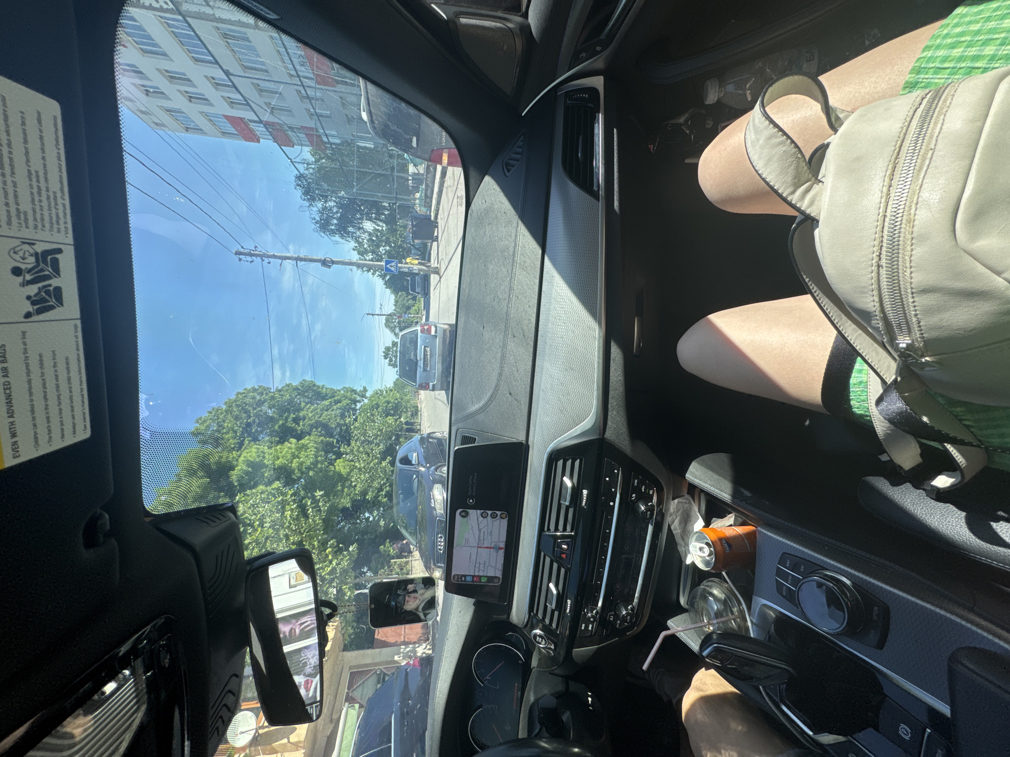 The image, taken from inside a car, shows an urban street scene in Byala Slatina, Bulgaria. The street includes buildings, trees, telephone poles, and parked cars. Inside the car, a backpack rests on the passenger's lap. On the car's dashboard, a can, a cup with a straw, and a phone displaying a map are visible. The car appears to be stopped in traffic.