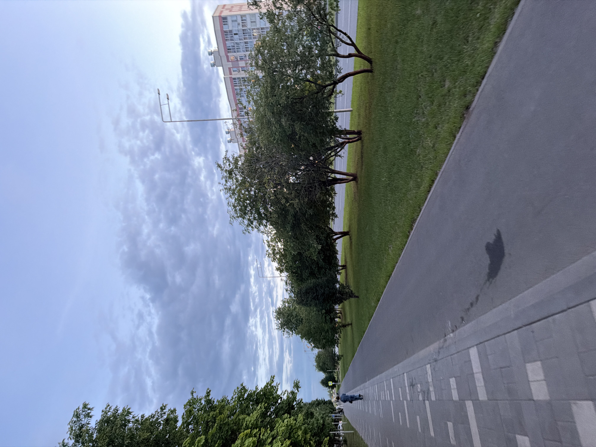 The image depicts a public walkway or bike path in Nizhnekamsk, Russia. The path is paved with asphalt, bordered by a strip of green grass and a decorative tiled pavement. Several trees with sparse foliage are planted along the grassy area, and a multi-story building is visible in the background. The sky is partly cloudy, suggesting it could be daytime with variable weather.
A person is seen on the pathway, appearing to be rollerblading or skateboarding, indicated by their posture and the blur of motion. The overall scene is one of urban activity, with infrastructure like streetlights and traffic signals hinting at a developed area. The time of day is unclear, but the lighting suggests it is not the peak of midday sun. The presence of trees and grass implies an outdoor setting, possibly a park or a residential area with green spaces. The building in the background, with its numerous windows, suggests it is an apartment complex or office building.
