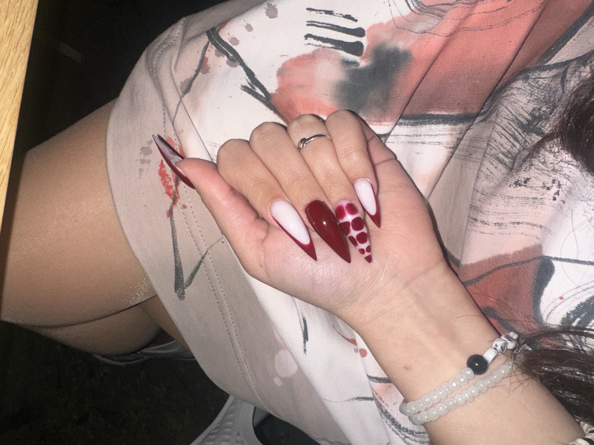 A high-angle shot depicts a person's hand and arm adorned with jewelry and elaborately decorated nails. The nails are long and pointed, displaying a mix of white tips, solid red, and red-and-white polka dot designs. A simple silver ring is on the ring finger. A beaded bracelet is around the wrist. The person's arm rests on a surface covered with a patterned fabric. Part of the leg, sheathed in a shiny stocking material, is visible in the background. No information suggests the location is Yerevan, Armenia.
