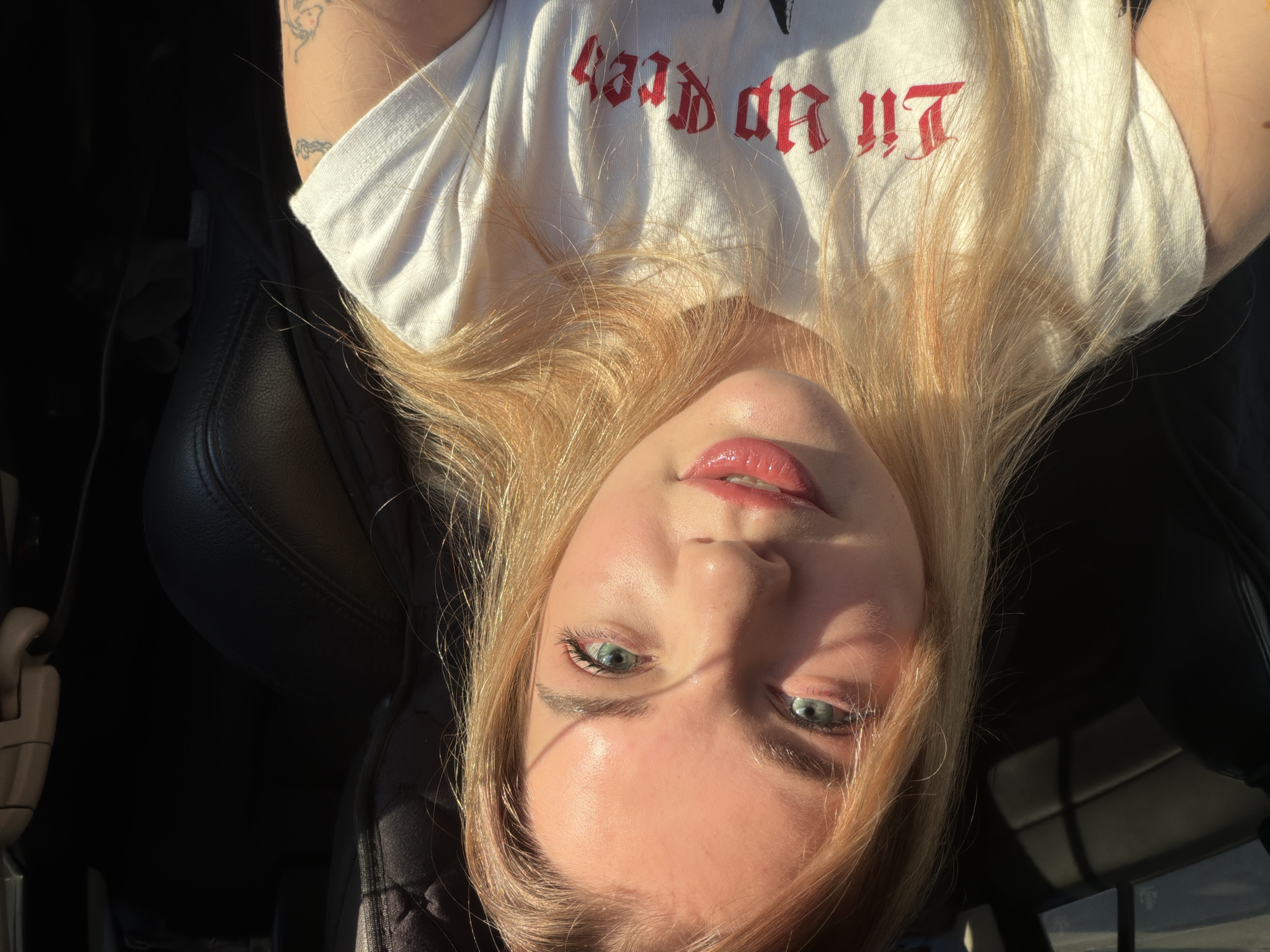 The image is a selfie of a young, light-skinned woman with blonde hair and blue eyes. She is wearing a white t-shirt with red gothic-style lettering that reads "Lil Peep". The photo is taken from a low angle, looking up at her face. The background shows a black leather car seat and some glimpses of the outside, possibly through a car window. While I cannot pinpoint the exact location within Naberezhnye Chelny, Russia, the context suggests she is inside a car, possibly parked or driving.