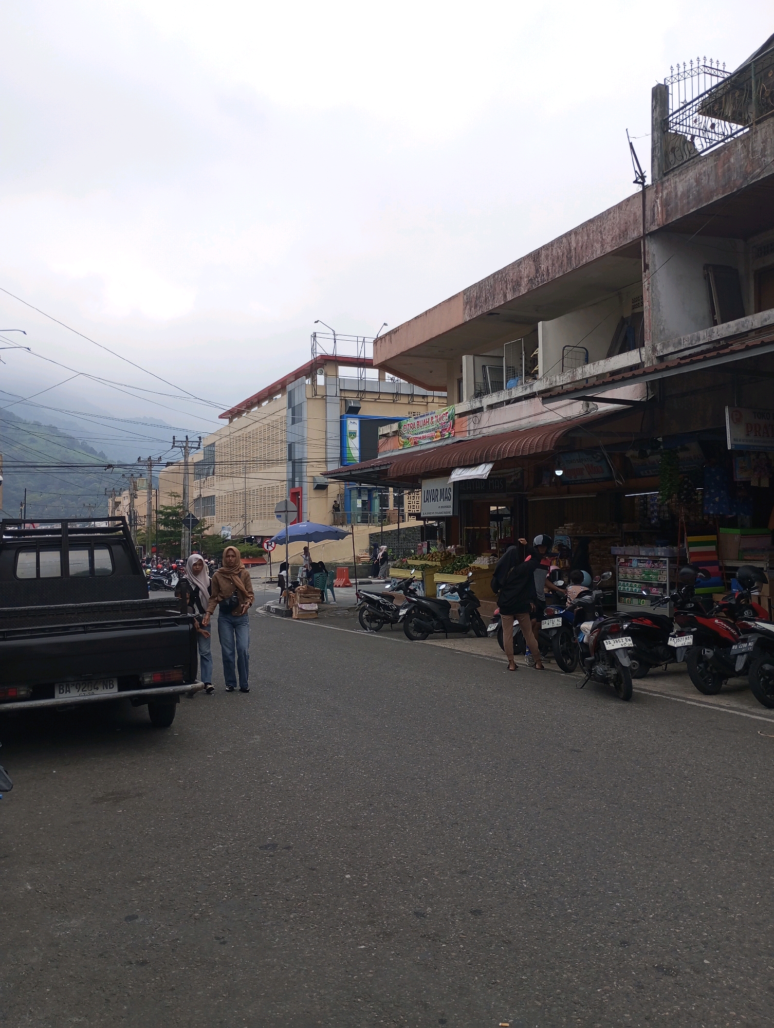 The image depicts a street scene in Padang Panjang, Indonesia, likely a commercial area given the presence of shops and the variety of parked motorcycles. In the foreground on the left, a black pickup truck is parked, its rear end visible. Further down the street, two women wearing hijabs are walking on the asphalt road. The woman on the left wears a light-colored hijab and a dark top with jeans, while the woman on the right wears a brown hijab and a matching brown jacket over jeans, carrying a camera.
Along the right side of the street, a row of shops is visible. One shop has a sign that reads "LAYAR MAS". Another sign above a storefront appears to advertise "CITRA BUAH & JUICE". Several motorcycles are parked neatly along the sidewalk in front of the shops, suggesting a busy commercial hub. A blue umbrella is open near the shops, and a few people are seated or standing nearby, possibly customers or vendors. In the background, a multi-story building with a distinctive facade and a large billboard is visible, and beyond that, rolling hills under an overcast sky. The overall atmosphere is that of an active urban environment with elements of everyday commerce.