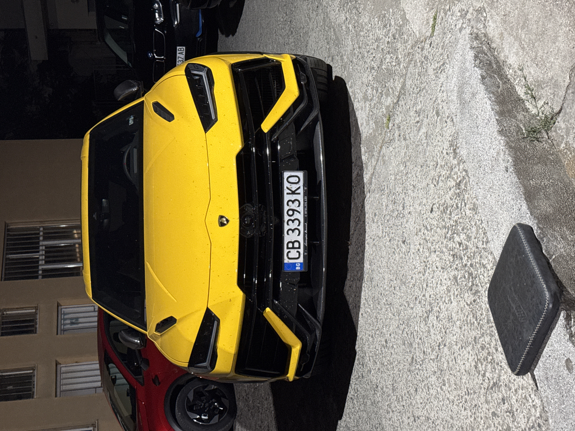 In Burgas, Bulgaria, a bright yellow Lamborghini Urus is parked on a concrete surface. The vehicle's front is facing the camera, showcasing its distinctive headlights, grille, and license plate which reads "CB 3393 KO". To the left, a red vehicle is partially visible, and to the right, a black vehicle with a partial license plate "37 AB" is parked. Behind the cars, a building with windows featuring metal bars is visible. The overall scene is dimly lit, suggesting it is either nighttime or dusk. A small, black, square object with white stitching sits on the ground in the foreground.
