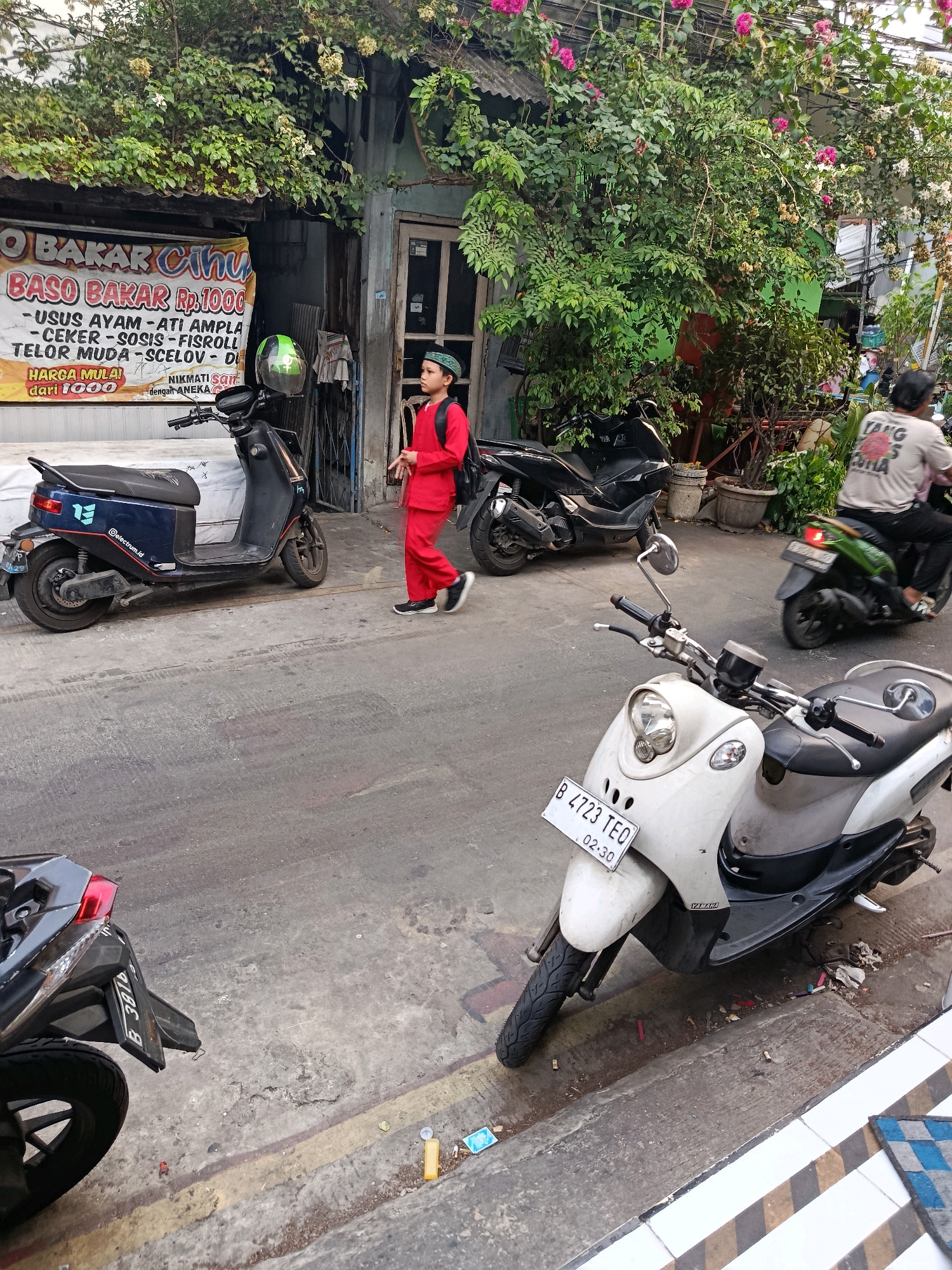 In a street scene in North Jakarta, Indonesia, a young boy wearing a red outfit and a peci hat walks down the sidewalk next to a building with a sign advertising "BASO BAKAR Rp. 1000". He carries a backpack. Several motorcycles are parked and in motion. A dark blue electric scooter is parked on the left. A white scooter with license plate B 4723 TEQ is in the foreground. Behind the boy, a black motorcycle is parked. Further in the background, a person on a green scooter rides away from the camera. The building facade features a window, greenery, and overhead vines with pink flowers. The street is paved with concrete and asphalt, with scattered debris and yellow lines marking the edge of the sidewalk.
