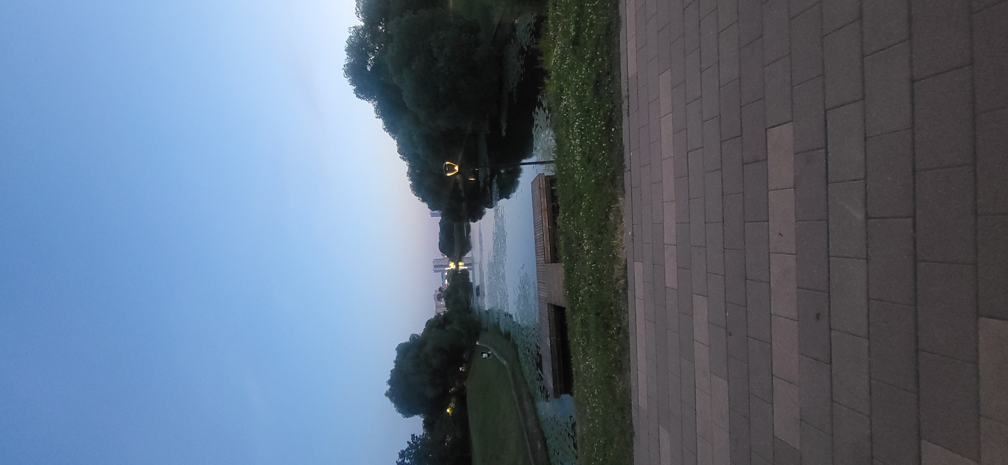 A vertical, full-shot image captures a park at dusk with a canal or narrow river running through its center, reflecting the twilight sky. The foreground is dominated by a paved walkway made of rectangular stone pavers in varying shades of gray and brown. Beyond the walkway, a grassy area with scattered small white flowers leads to the water. A wooden pier or dock juts out into the water from the left bank.
The water itself is partially covered with lily pads and other aquatic vegetation, creating a textured surface. A single lamppost with an illuminated, ornate fixture stands on the right bank of the water. In the middle ground, lush green trees with full canopies line both sides of the water body. Distant buildings, appearing as light-colored high-rises, are visible on the horizon, suggesting an urban setting. Two indistinct figures are walking on the grassy bank on the left side of the canal. The sky is a clear, gradient blue, transitioning to a lighter hue near the horizon, indicating the end of daylight. The overall impression is of a tranquil evening scene in a city park.