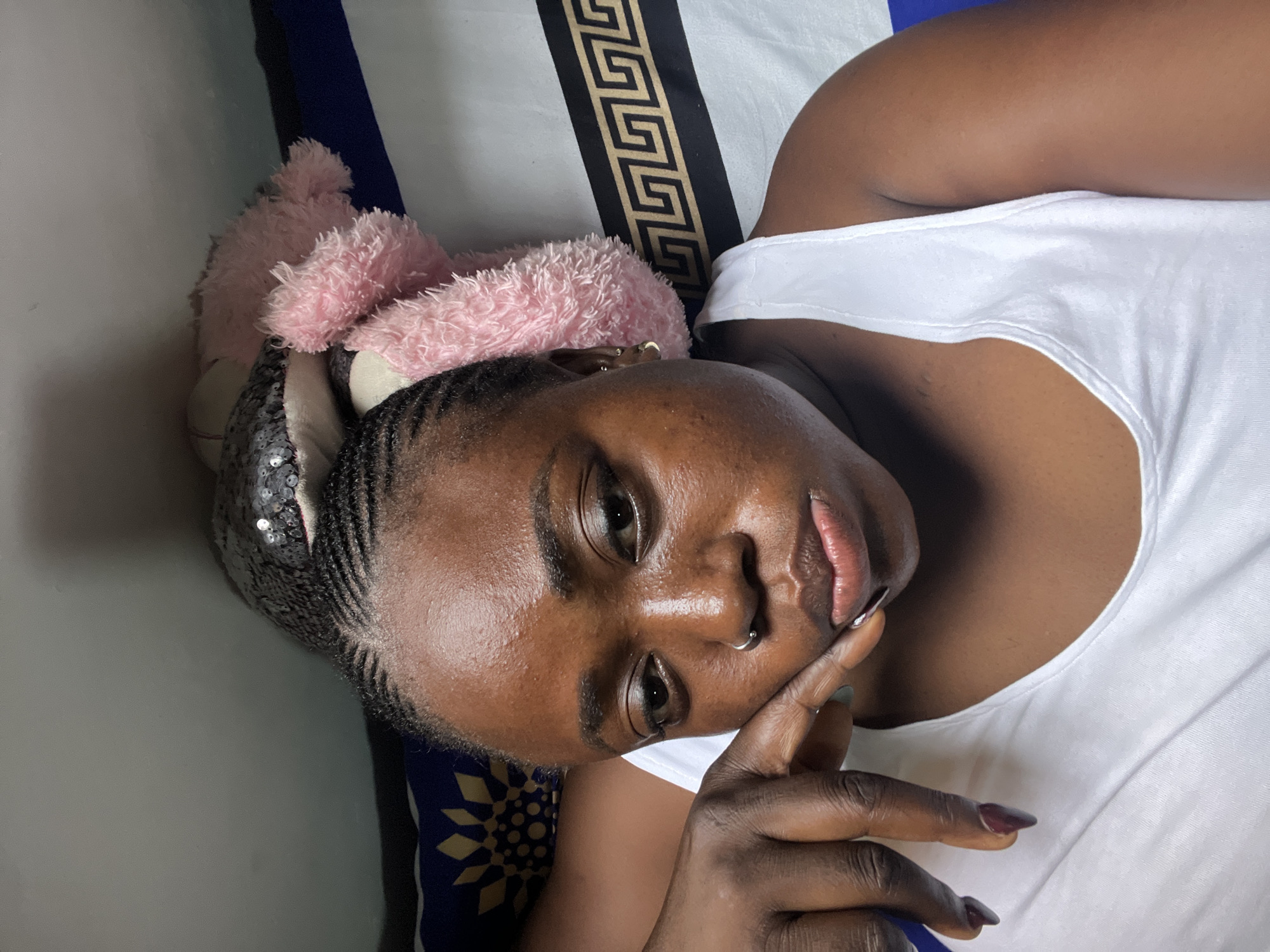 A selfie of a Black woman lying on a bed, with her head resting on a stuffed toy. She is wearing a white tank top and has her hair braided. The background shows bedding with a patterned design. The scene appears to be indoors. There is no information in the image that can confirm the location as Abuja, Nigeria.
