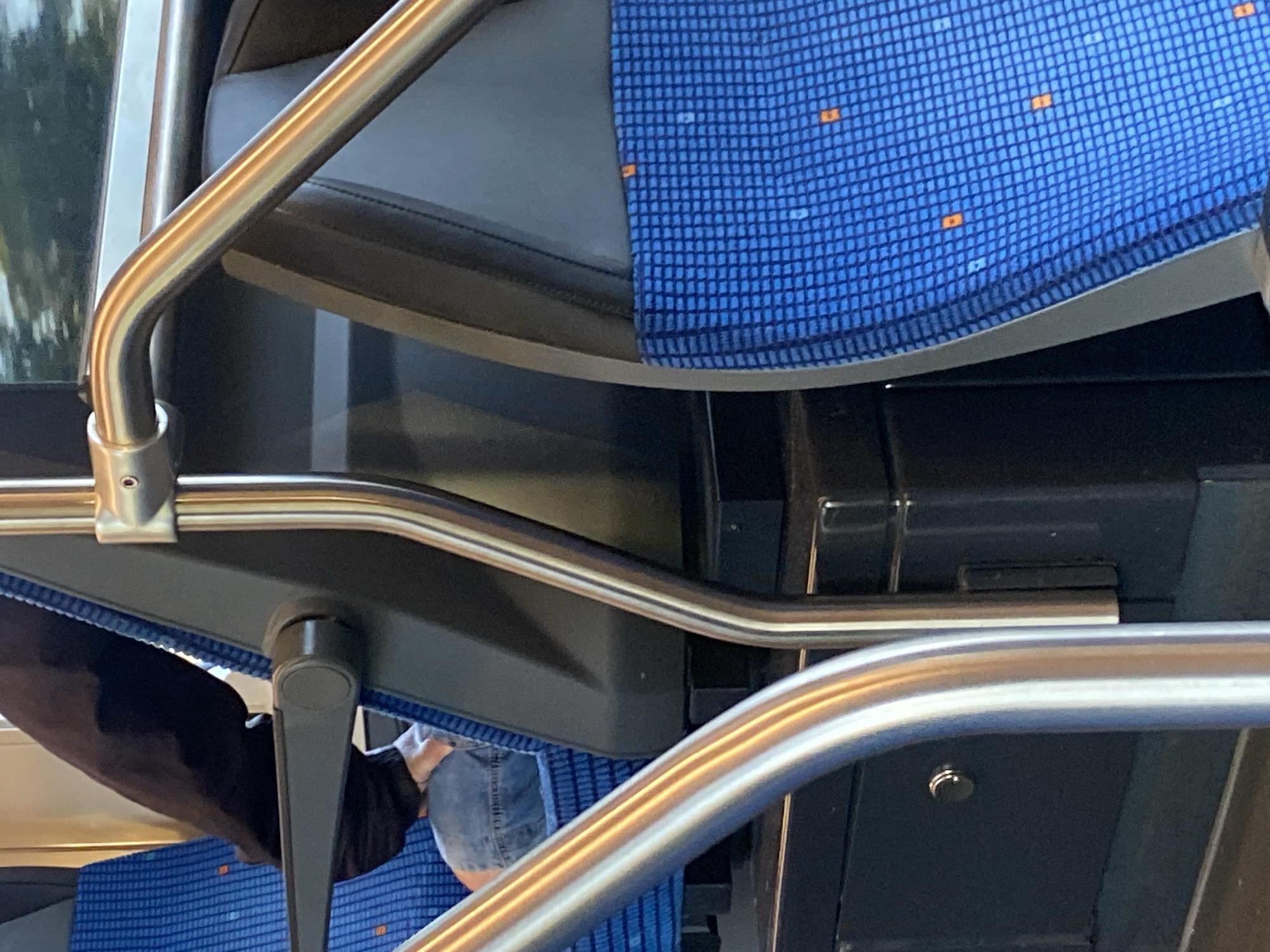 The image shows the interior of a public transport vehicle, likely a bus or train, with blue patterned seating and silver grab rails. A person is partially visible, wearing blue jeans and a dark jacket, seated on the blue patterned seat. The environment appears to be indoors. The city of České Zlatníky, Czechia, is a small village and not a city, so it is unlikely that this image is located there.