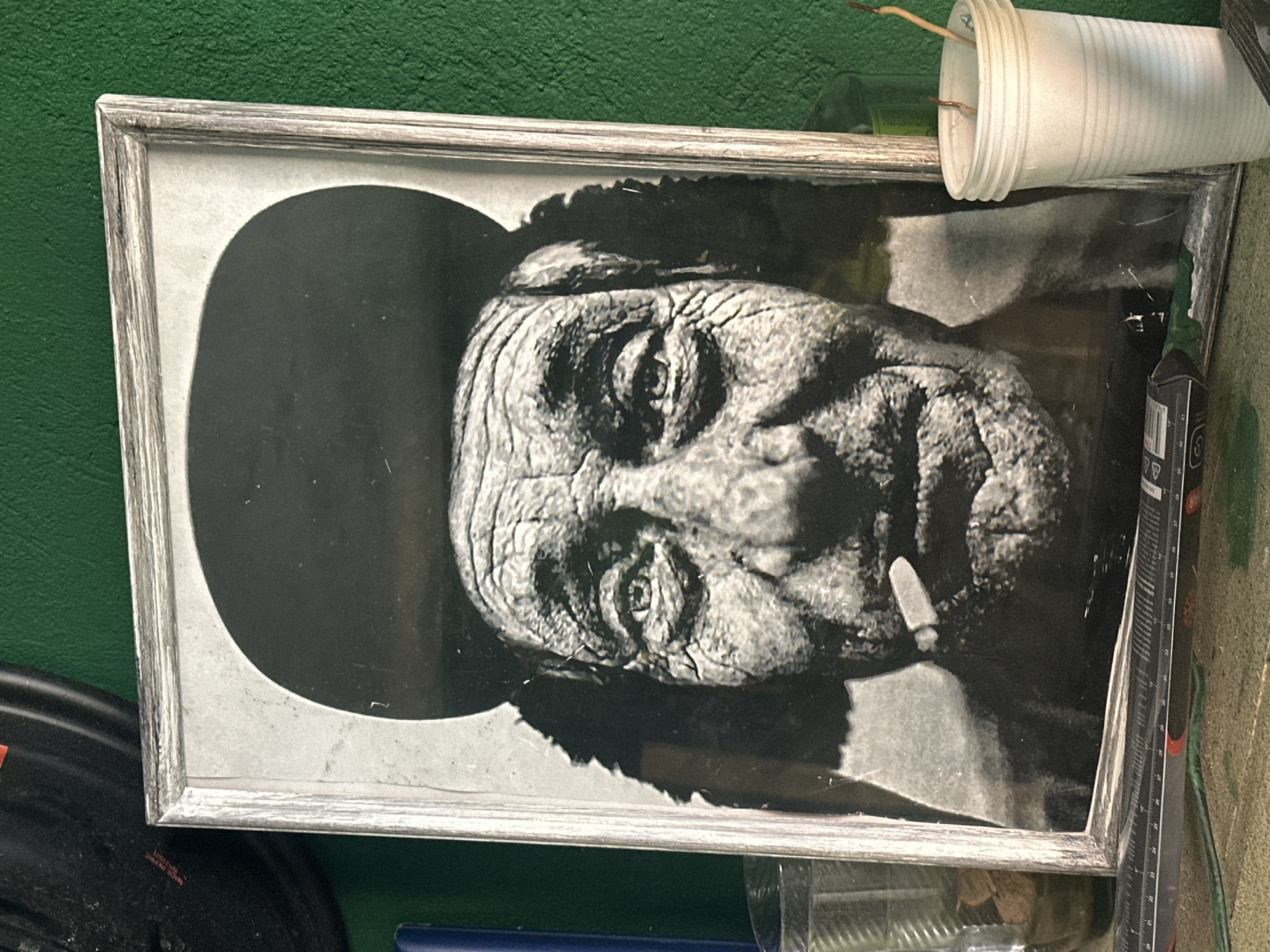The image shows a framed black and white portrait of an elderly man. The man is wearing a hat and smoking a cigarette. The frame is silver. The picture is located indoors in a cluttered environment, appearing to be a workshop or storage area. Debris and various objects surround the frame, including plastic cups, a ruler, and other unidentified items. The background wall is green. The location is not possible to confirm within Andreyevka, Russia, from the image.