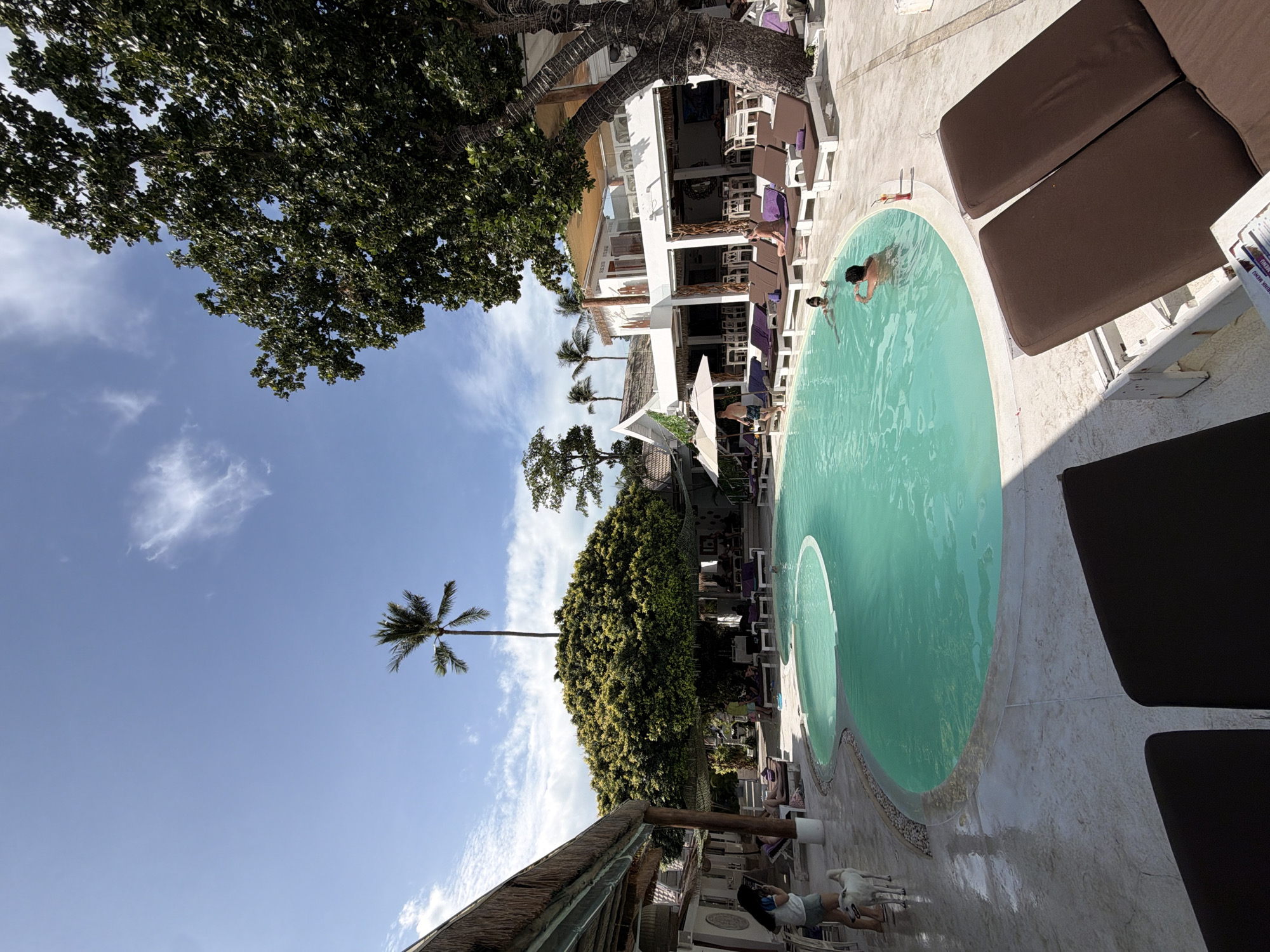 The image depicts a poolside scene at a resort or hotel in the Ko Samui District, Thailand. The setting appears to be a resort with a large, kidney-shaped swimming pool filled with clear turquoise water. Around the pool, there are numerous lounge chairs with brown cushions, some occupied by people relaxing.
Several individuals are visible in and around the pool. Two people are swimming in the pool, one appearing to be doing a breaststroke while the other is submerged. Another person is standing at the edge of the pool, facing away from the camera. A dog is also present near the pool, standing next to a woman holding a phone. In the background, there are buildings with thatched roofs and white facades, suggesting a tropical resort. Lush greenery, including a large, dense tree and palm trees, dominates the landscape, contributing to the tropical ambiance. The sky is blue with scattered white clouds. The overall impression is one of a relaxed, sunny day at a tropical resort.