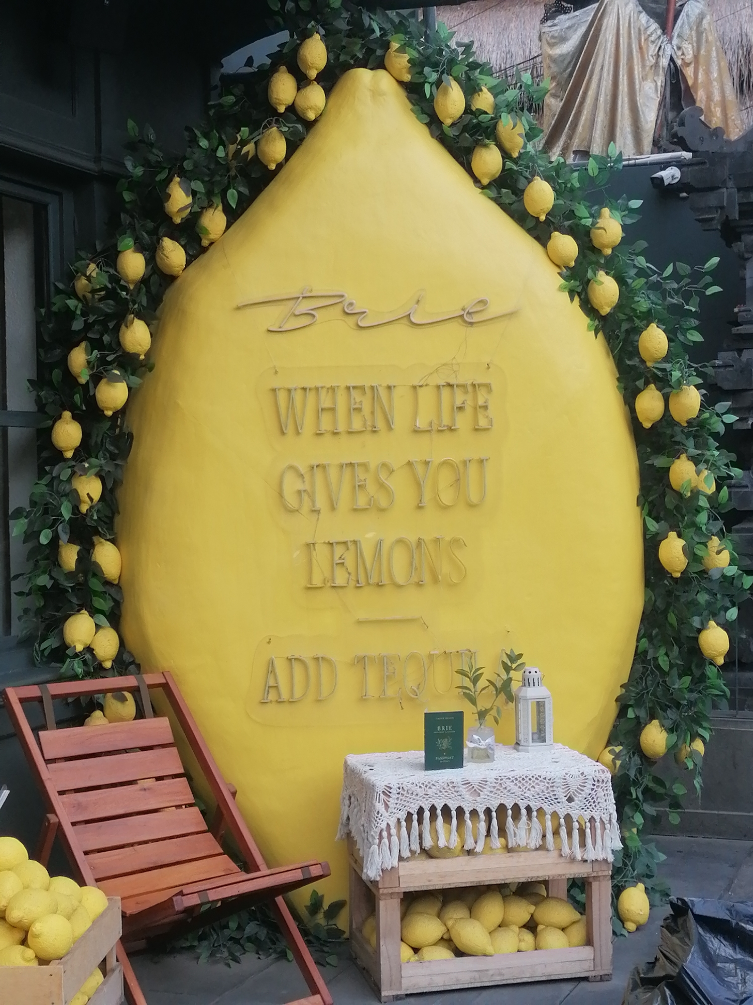 A bright yellow, oversized lemon structure serves as a backdrop in what appears to be an outdoor seating area. The structure is decorated with a vine of artificial lemons and green leaves. Inscribed on the lemon structure are the words "Brie," "WHEN LIFE GIVES YOU LEMONS," and "ADD TEQUILA." In front of the lemon, a wooden deck chair is placed at an angle. To the right of the chair, a small table is draped with a white crocheted cloth and adorned with a small green book, a glass vase with greenery, and a white lantern. Beneath the table, a wooden crate is filled with lemons. To the left of the chair, another wooden crate is partially visible, also containing lemons. The overall scene suggests a themed decoration, likely for a cafe or restaurant. The location is identified as Ubud District, Indonesia.