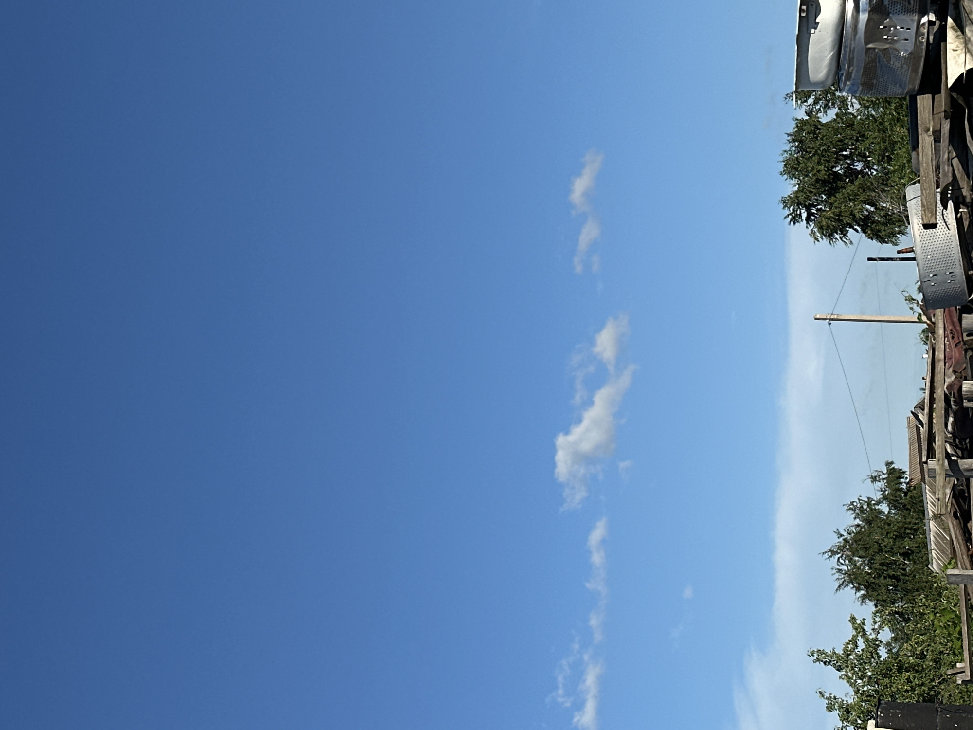 The media file shows a bright, clear blue sky with a few wispy white clouds on the right side. The image is oriented vertically, with the sky occupying the majority of the frame. On the far right, there is a section of what appears to be an outdoor junkyard or storage area. This area includes wooden structures, metal objects that resemble parts of appliances, and lush green trees. A wooden utility pole with wires is also visible, suggesting an inhabited area. The lighting indicates it is daytime, likely mid-morning to afternoon, given the brightness and the clear sky. There are no people or animals visible. The overall impression is one of an outdoor, possibly rural or semi-industrial setting on a sunny day. There is no visible text in the image. The location context provided is Tobyl, Kazakhstan.