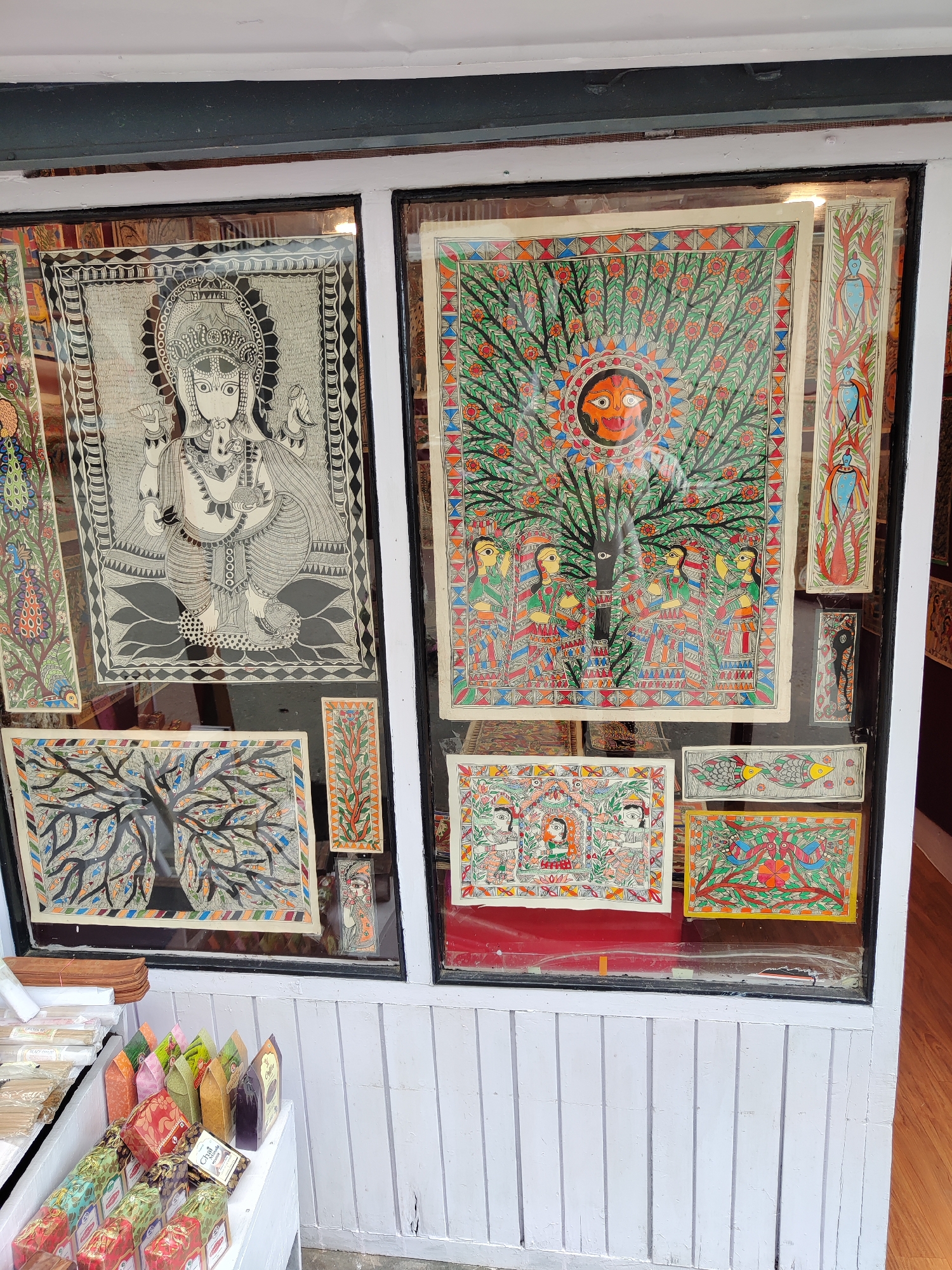 The image displays a vibrant storefront window showcasing a collection of traditional Indian folk art, primarily Madhubani paintings. The scene suggests a shop or gallery specializing in cultural handicrafts.
The window is divided into two main sections by a white vertical frame. In the left section, a prominent large black and white painting depicts the Hindu deity Ganesha, seated and adorned with intricate patterns and jewelry, framed by a geometric border. Below this, a smaller rectangular painting features a stylized "tree of life" with fish integrated into its branches, rendered in muted colors. A vertical painting with colorful peacocks and floral motifs is partially visible to the left.
The right section of the display is filled with an array of colorful artworks. A large central painting depicts a vivid "tree of life" with a radiant, orange sun-like face at its top, surrounded by green foliage and red blossoms. Several female figures are shown interacting with the tree. To its right, several smaller, vertical paintings feature stylized birds and abstract designs. Below the large tree painting, two more rectangular pieces are displayed: one with human figures amidst floral patterns, and another featuring two colorful birds perched on a branch.
Below the window, on a white wooden shelf, various retail products are arranged. These include numerous small, brightly colored boxes, some triangular, and other items wrapped in clear plastic, possibly indicating incense or other small gifts. One dark box on the shelf clearly shows the text "Chai" and "Original," suggesting a tea product. The white wooden paneling below the window adds to the traditional shop aesthetic. The time of day appears to be daylight, as indicated by the general illumination.