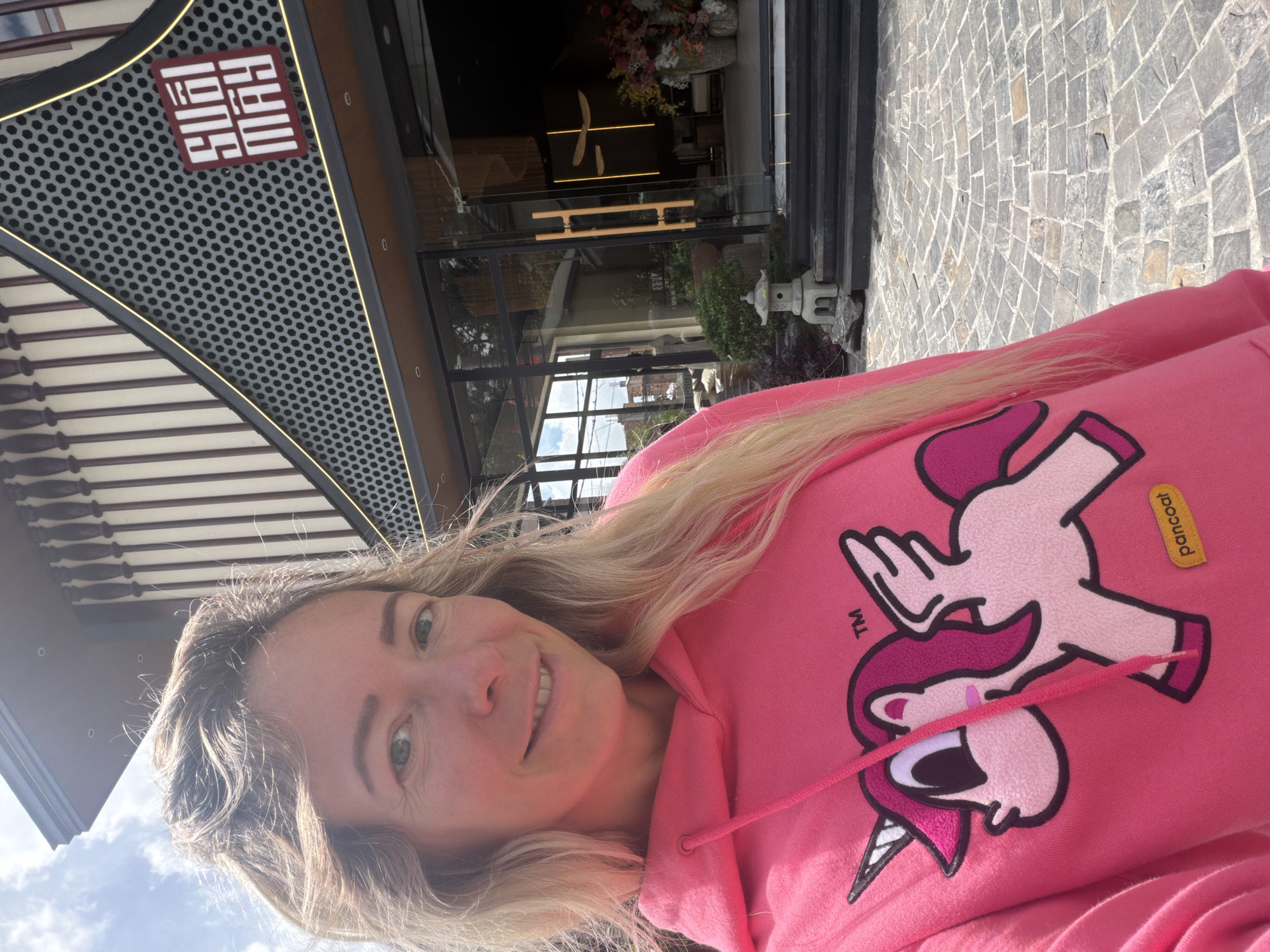 A woman with blonde hair is taking a selfie outdoors in front of a building. She is wearing a bright pink hoodie with a cartoon unicorn design. The building appears to be a restaurant or shop with a modern architectural style. Above the entrance, there is a sign with the words "SUAL MAY" in a distinctive font. The ground in front of the building is paved with stone. The scene suggests a bright, sunny day. The location is Da Lat, Vietnam.