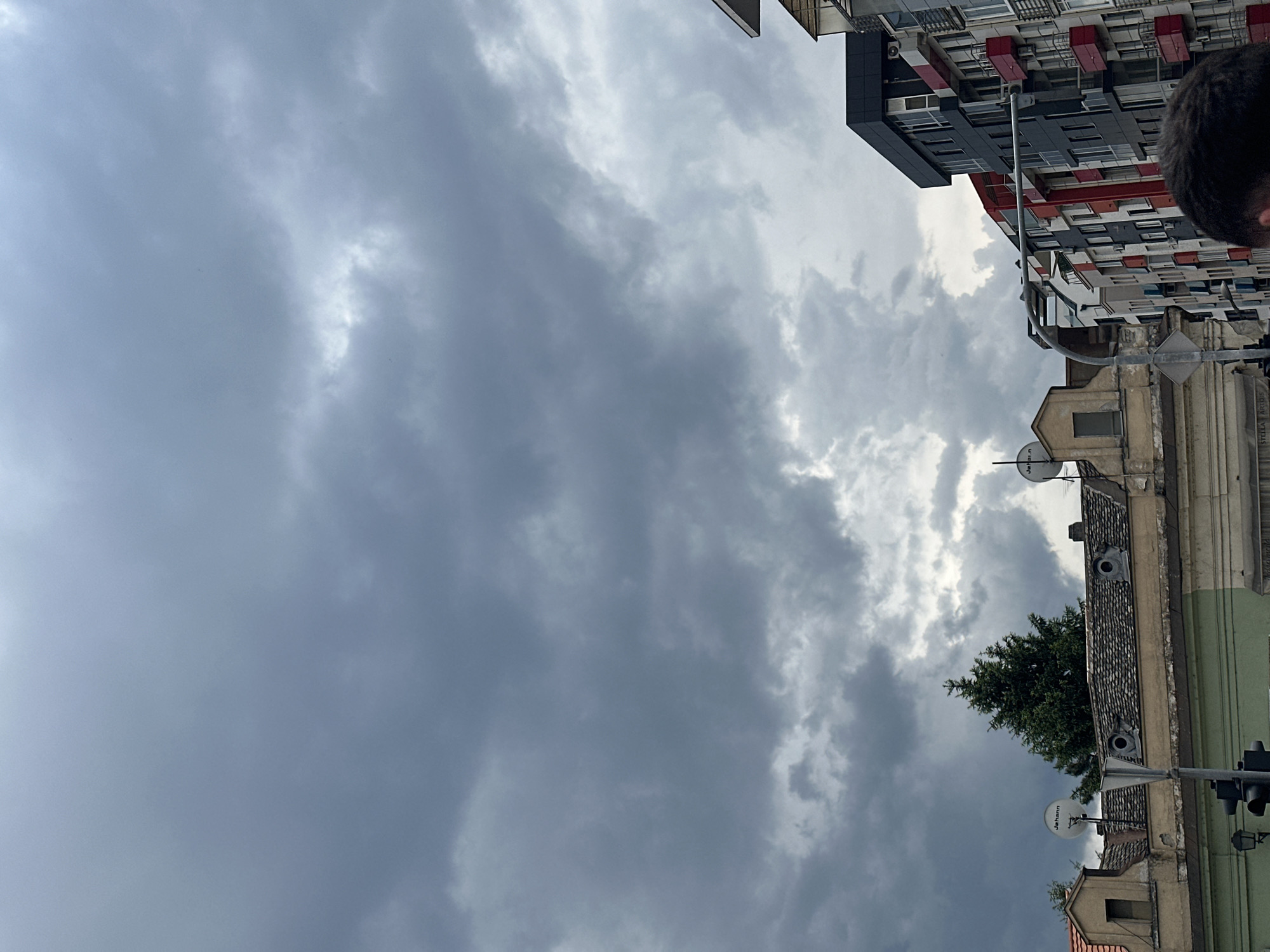 The image shows a cloudy sky over a street in Novi Sad, Serbia. A tall, modern building with red and grey accents is visible on the right side of the frame. Below the modern building, there is an older building with a tiled roof, satellite dishes, and a green facade. A tree is positioned between the old buildings. A traffic light is on the right side, next to the building. The clouds are dark and cover most of the sky. A portion of a person's head is visible on the right side of the image.