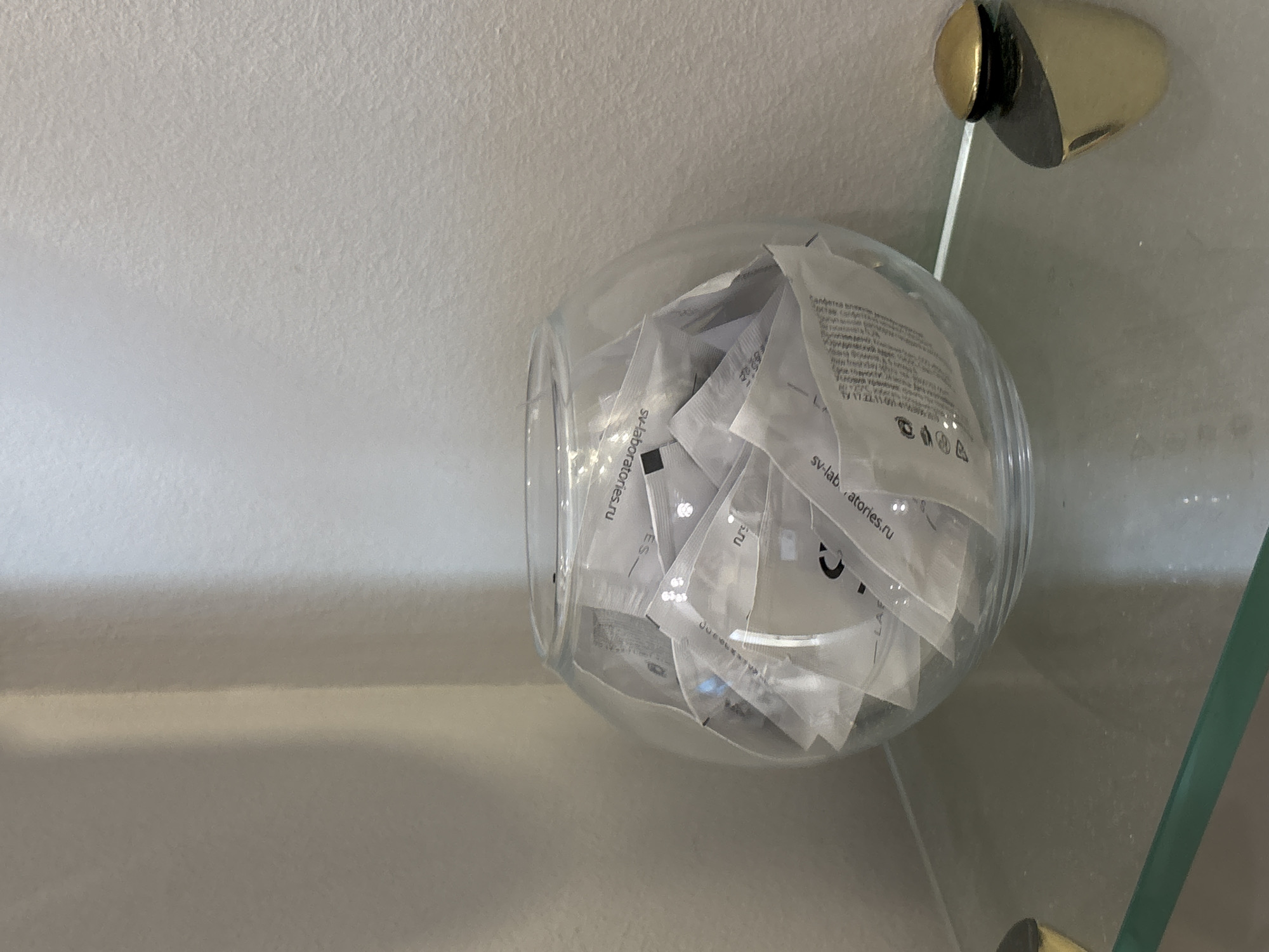 A clear glass bowl is filled with many small, white, individually wrapped packets. The packets have text and logos on them. Some of the text visible includes "sv-lab.atorles.ru". The bowl sits on a shelf. To the right of the bowl, a glass shelf is visible, supported by a gold fixture attached to a white wall. The lighting appears to be artificial, casting a soft illumination on the scene. The overall impression is one of organization and perhaps storage of samples or small products. The environment is clean and uncluttered.
