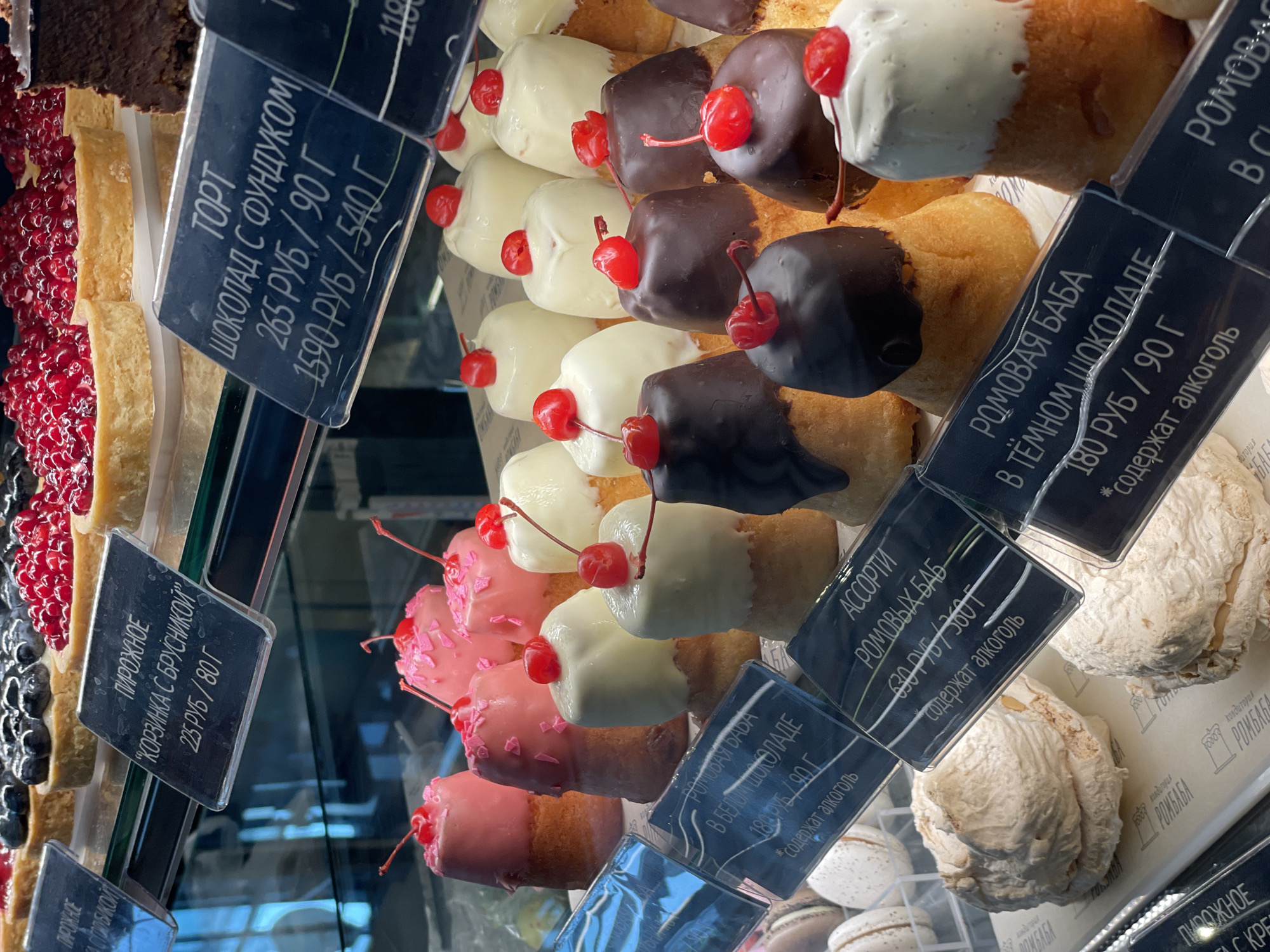 The image displays a close-up view of a bakery display case filled with various pastries. In the foreground, there are two large meringue-like desserts. Behind them, a row of small cakes topped with cherries is visible, some with white icing and others with dark chocolate icing. Further back, there are pink-frosted pastries, also decorated with cherries. In the background, several tarts are visible, some topped with red berries and others with blueberries. Price tags are placed in front of the desserts, with handwritten text in Russian indicating the names and prices of the items. The environment suggests a retail setting, likely a patisserie or bakery. There are no people visible in the image. The image is a high-angle shot looking down into the display case. The overall impression is one of a well-stocked and appealing dessert selection.