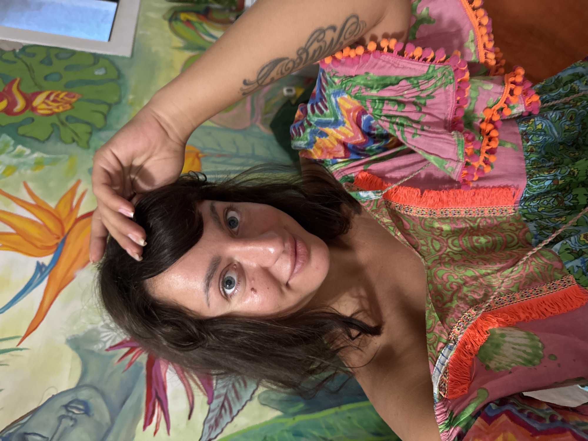 The image shows a woman with dark, wavy hair and blue eyes, wearing a colorful off-the-shoulder dress. She is touching her hair with her right hand. The background is a mural with tropical plants and a figure with blue skin. The image was taken in San Benedetto del Tronto, Italy.