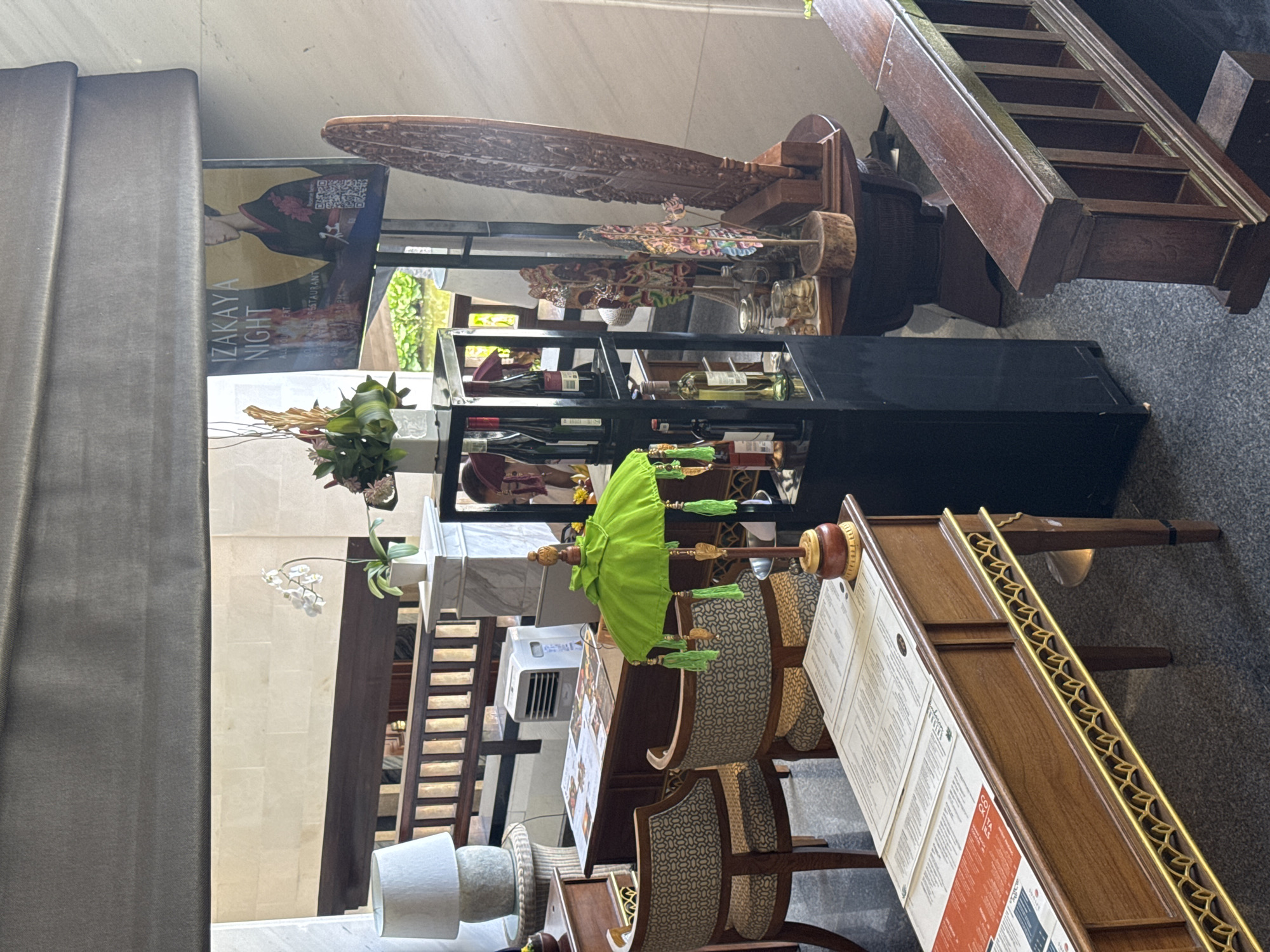 The image depicts the interior of what appears to be a restaurant or hotel lobby in Kuta Selatan, Indonesia, with a focus on a decorated display area. In the foreground, a wooden table with intricate gold detailing displays several menus. To the right, a dark wooden bench with open cubbies is visible. Behind the table, two upholstered chairs with a patterned fabric face towards the display. Dominating the center is a tall, black shelving unit holding bottles of wine and other decorative items. A bright green, tasseled umbrella-like decoration sits on a wooden base on this shelf. To the left of the shelving unit, a white air conditioner unit is present next to a wooden desk with more menus. Further to the left, a tall lamp with a white shade rests on a woven stand. In the background, a large screen displays an advertisement for "IZAKAYA NIGHT" featuring a person in traditional Japanese attire. Various decorative elements, including wooden carvings and a flower arrangement, are scattered throughout the scene, suggesting a curated aesthetic. The environment is well-lit, with natural light filtering in from an unseen source.