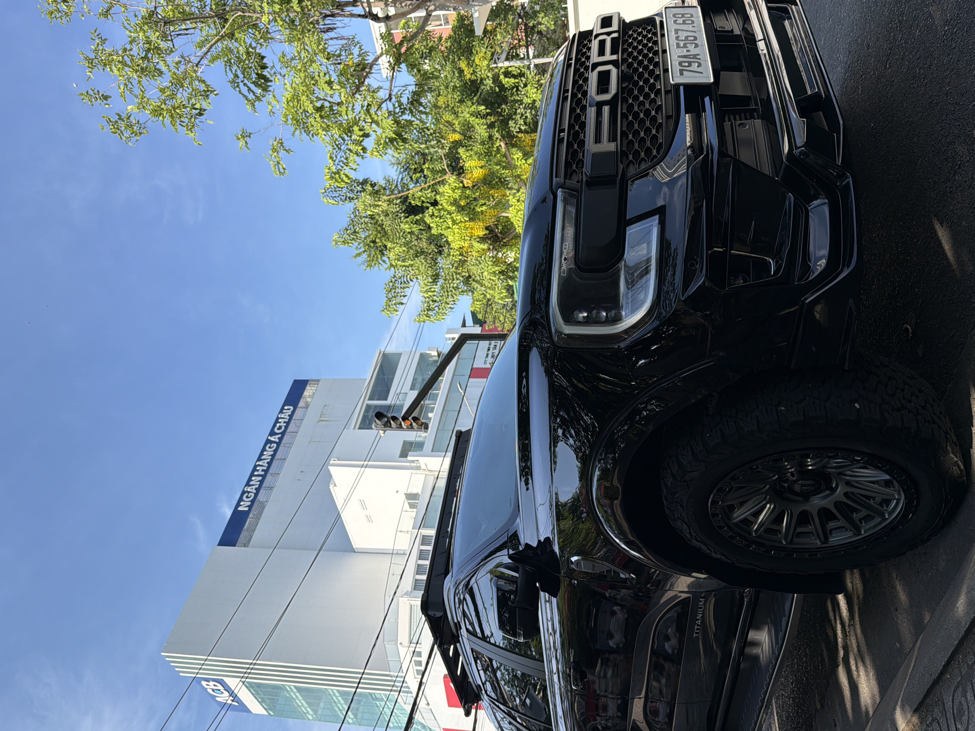 The image captures a black Ford Everest parked on a city street in Nha Trang, Vietnam, during daylight. The vehicle, featuring a roof rack and a prominent "FORD" grille, is positioned in the foreground, angled towards the viewer. Behind the car, a white building with blue signage that reads "ACB" and "NGÂN HÀNG Á CHÂU" stands tall, indicating it is a bank. Lush green trees with yellow blossoms flank the building and extend towards the clear blue sky. A traffic light is visible in the mid-ground, and power lines crisscross the scene. The street surface is asphalt, with shadows cast by the trees and the vehicle. No people are discernible in the image. The license plate of the Ford Everest reads "79A-567.68".