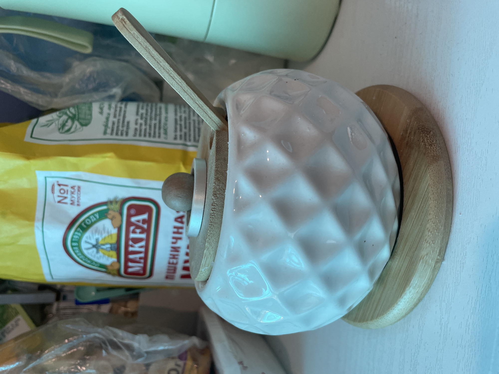 The image shows a kitchen setting in Olginka, Russia. A bag of Makfa brand wheat flour sits on a shelf along with other items packaged in plastic. A white ceramic sugar bowl with a textured surface, wooden lid and spoon rests on a circular wooden base. A green appliance sits in the upper background.