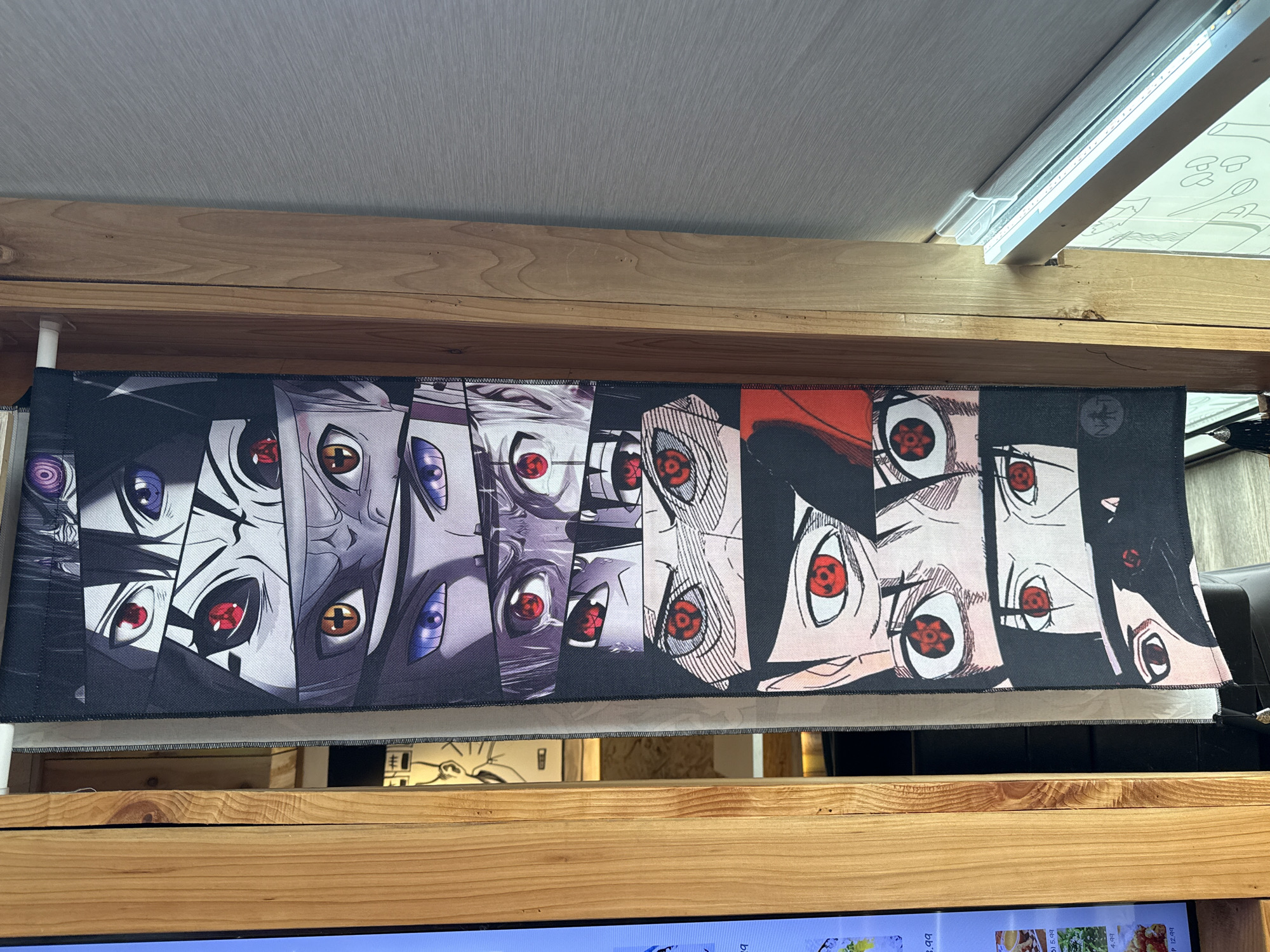A vertical tapestry featuring a montage of anime-style eyes hangs from a white rod, mounted on a wooden frame. The tapestry displays multiple pairs of eyes with distinct pupil patterns and colors, including red, purple, and orange. The eyes appear to be from various characters, likely from the anime series Naruto, given the characteristic red Sharingan and Rinnegan patterns. The tapestry is black with colorful eye illustrations. The wooden frame is to the left of the tapestry, and a gray textured wall is to the right. A portion of a digital menu board with food images and prices is visible on the far left, displaying items like "8.99" and "5.99". The background suggests an indoor setting, possibly a restaurant or a shop. The overall scene is well-lit, with natural light appearing to come from the right side. The location is identified as Tacoma, United States of America, but no specific street address or establishment is discernible.