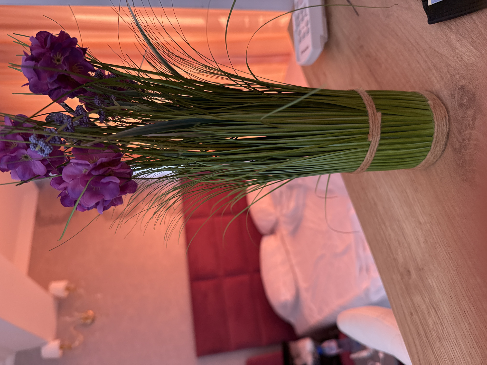 The image shows a close-up of a vase with artificial purple flowers and green grass on a wooden desk. In the background, a bed with white linens and a red headboard is visible. There are also wall lamps with white shades, a white chair, and curtains. The lighting suggests an indoor setting, possibly a hotel room. The city of Nesebar, Bulgaria, is not depicted in the image.